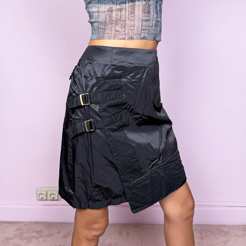 This vintage 2000s black high-waisted mini skirt features a structured wrap-over layer secured with buttons and adjustable straps, creating an asymmetric silhouette. Made from a shiny polyester-cotton blend with no stretch, it channels deconstructed dystopian and futuristic goth energy, perfect for night-out or statement looks. Finished with a back zipper closure. Excellent vintage condition.