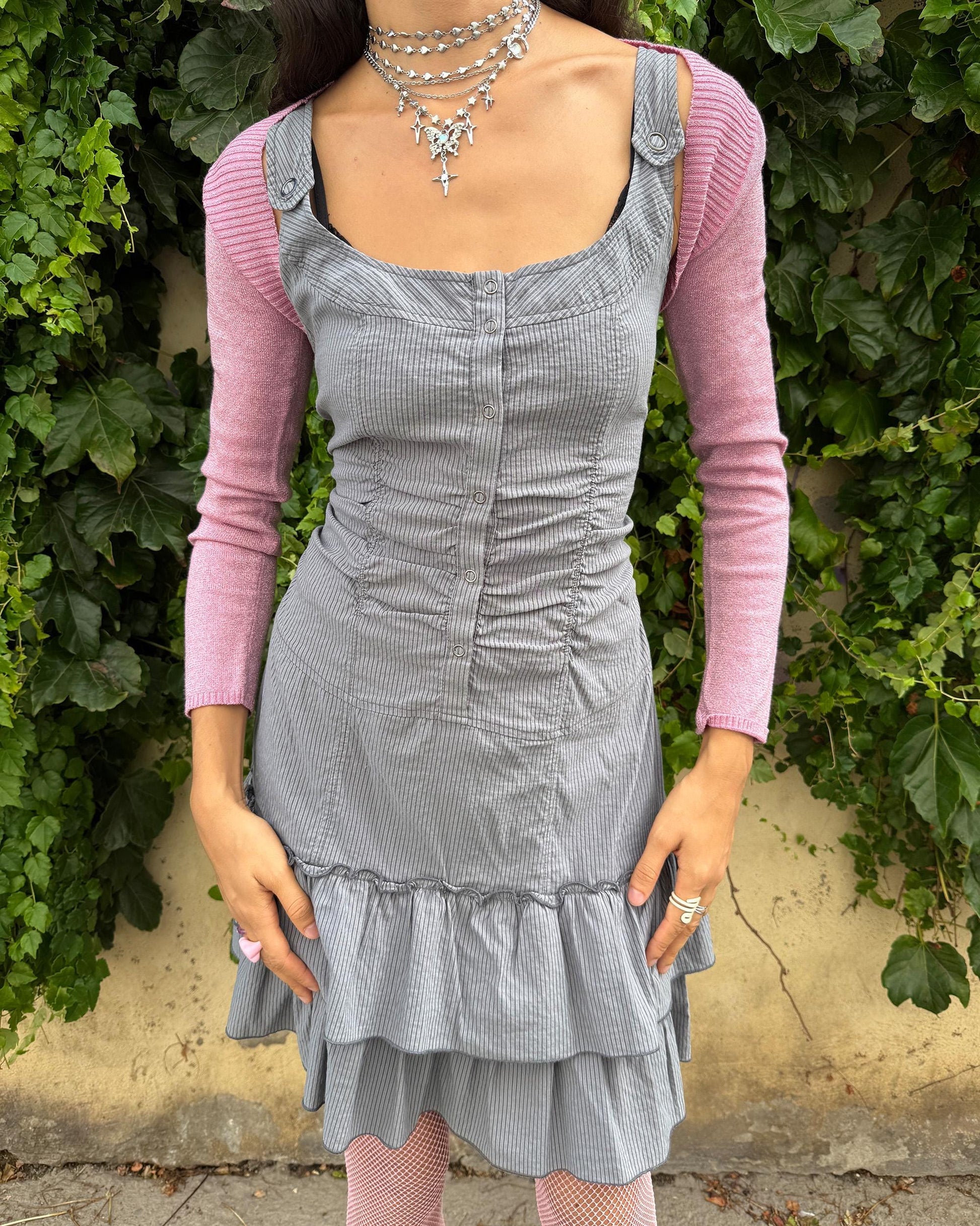 This vintage 2000s gray sleeveless mini dress features a striped pattern with ruched detailing, snap button closure, and a double-tiered ruffle hem. Its stretchy fit and structured silhouette evoke cyber-futuristic Harajuku streetwear and weird girl aesthetics, perfect for playful, statement layering. Made from a polyester blend. Excellent vintage condition.