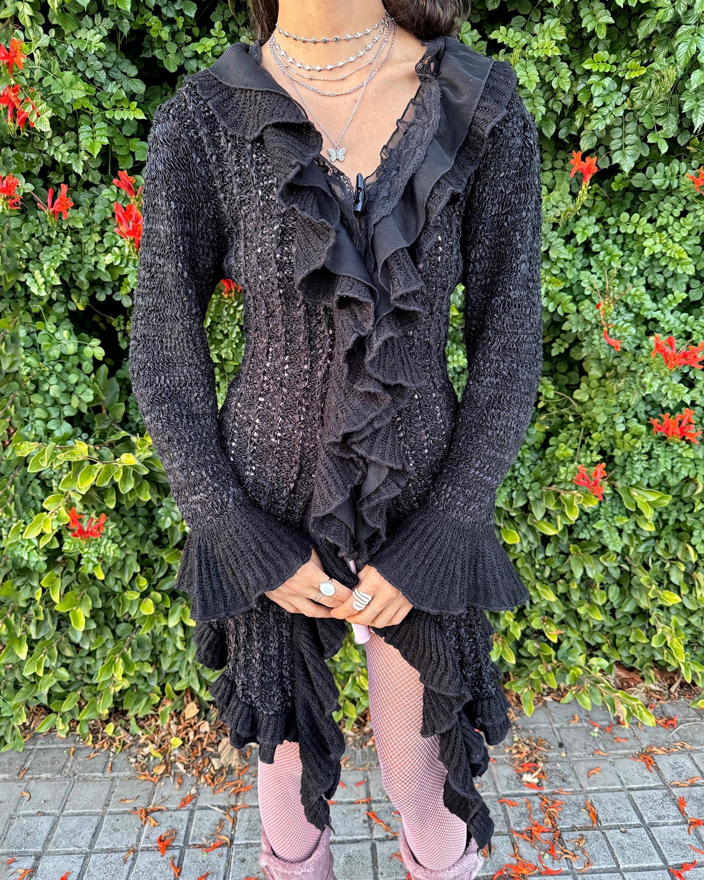 Vintage 2000s black longline cardigan featuring a dramatic ruffle neckline with lace details, bell-sleeve ruffle cuffs and a toggle front closure. The asymmetric pointy hem adds a whimsical, witchy silhouette, while the stretchy cotton–wool–polyamide blend creates a cozy, comfortable fit perfect for winter layering. A stunning fairy-goth statement knit jacket that blends elegance, softness and dark romantic style. Great vintage condition.