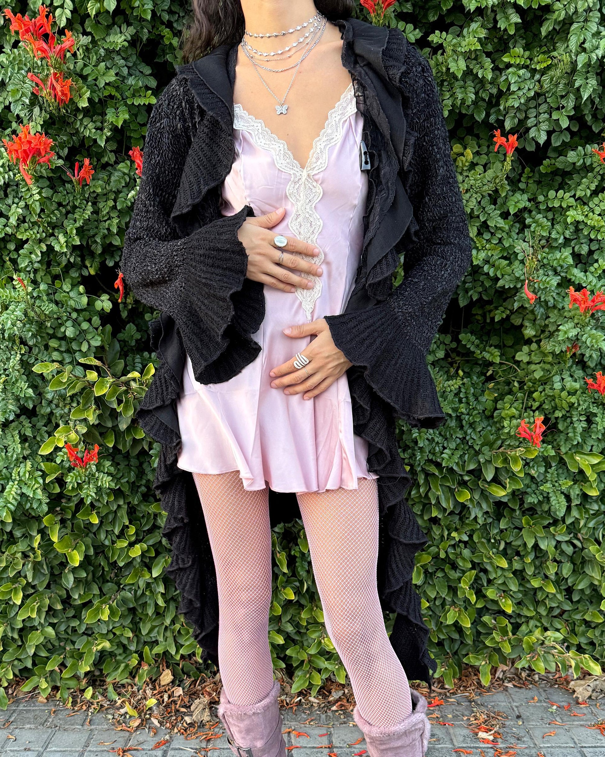 Vintage 2000s black longline cardigan featuring a dramatic ruffle neckline with lace details, bell-sleeve ruffle cuffs and a toggle front closure. The asymmetric pointy hem adds a whimsical, witchy silhouette, while the stretchy cotton–wool–polyamide blend creates a cozy, comfortable fit perfect for winter layering. A stunning fairy-goth statement knit jacket that blends elegance, softness and dark romantic style. Great vintage condition.