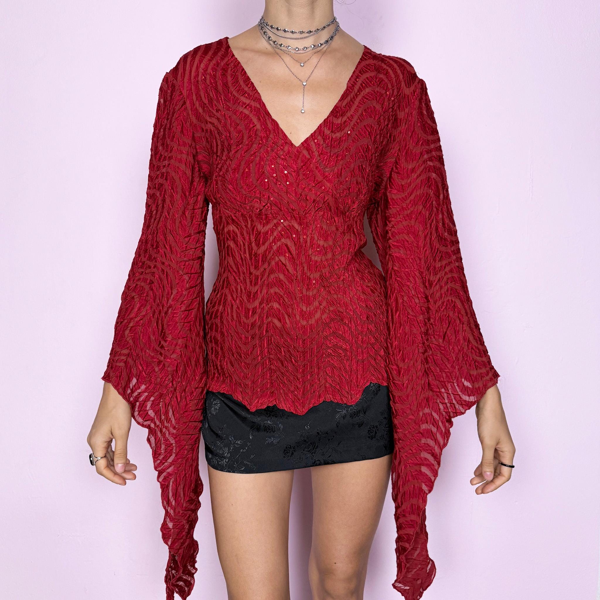This romantic glam vintage 2000s red blouse features a sheer burnout textured pattern with crinkled wavy detailing and subtle sequins. Designed with a wrap-style silhouette, three-quarter slit dramatic bell sleeves, and an asymmetric hem, it offers a sculpted yet fluid fit ideal for artistic parties, nights out, or editorial avant-garde styling. Made from stretchy polyester. Great vintage condition.