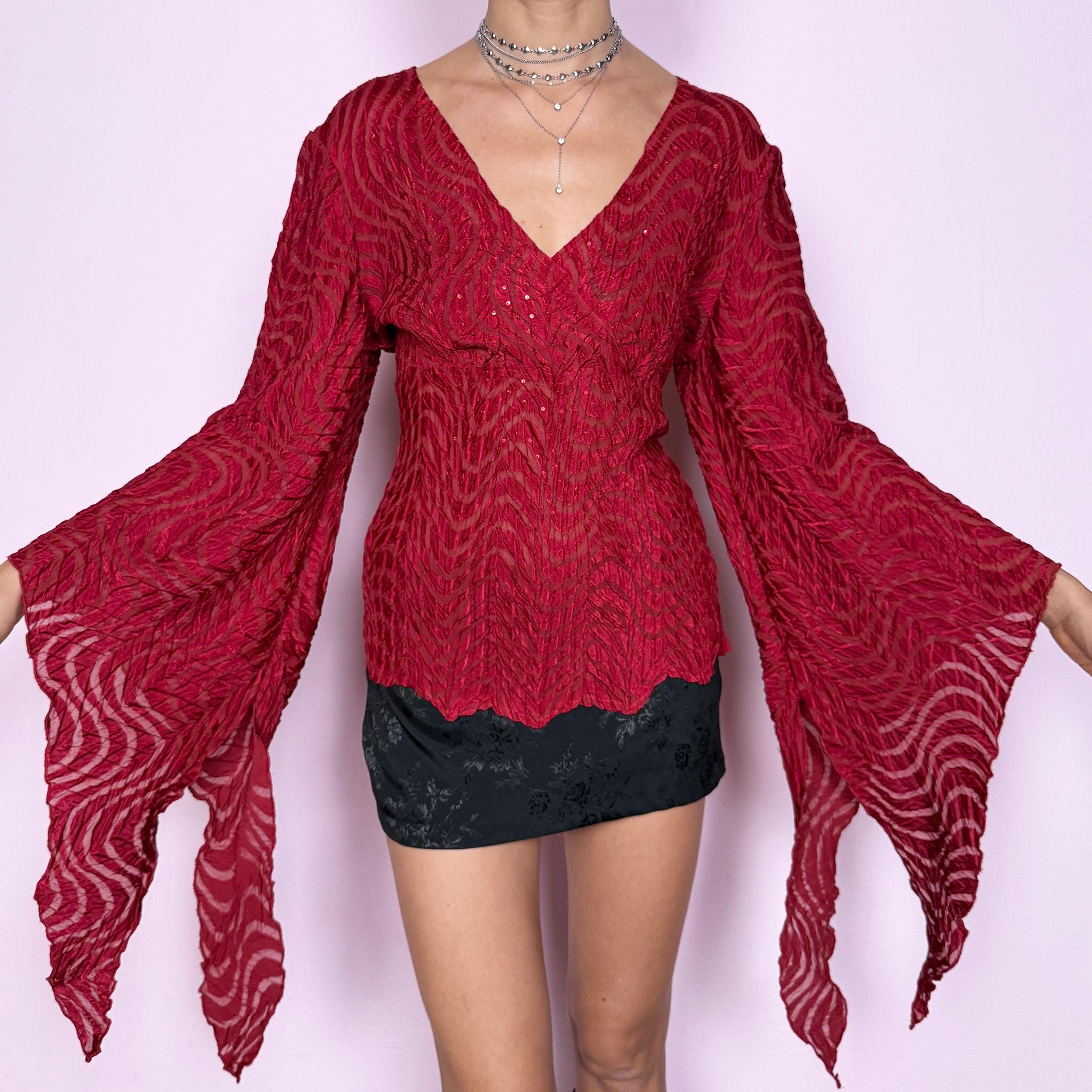 This romantic glam vintage 2000s red blouse features a sheer burnout textured pattern with crinkled wavy detailing and subtle sequins. Designed with a wrap-style silhouette, three-quarter slit dramatic bell sleeves, and an asymmetric hem, it offers a sculpted yet fluid fit ideal for artistic parties, nights out, or editorial avant-garde styling. Made from stretchy polyester. Great vintage condition.