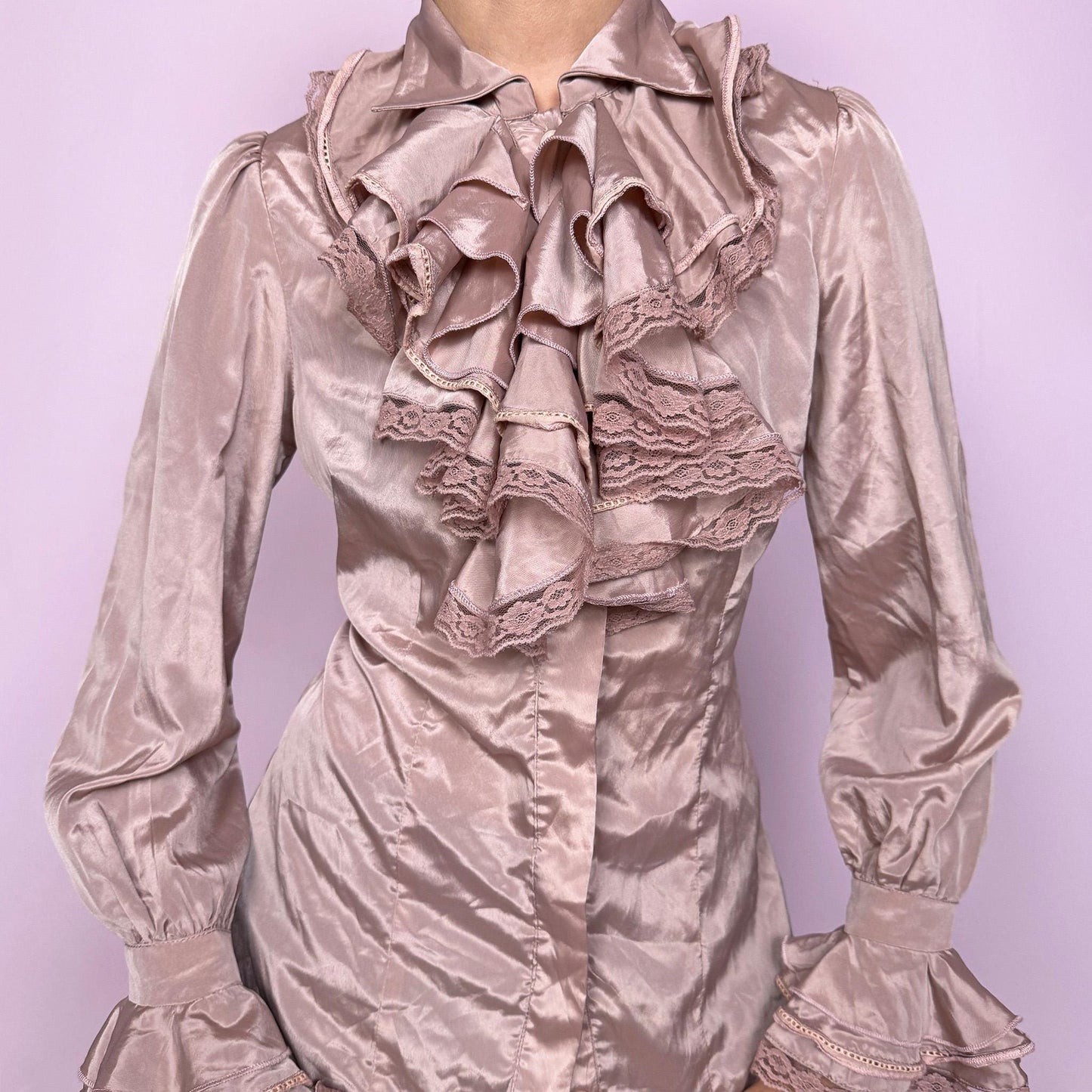 This romantic vintage 90s dusty pink shiny button-down collared blouse features dramatic exaggerated ruffles with lace trim and ruffle cuffs. The fitted, no-stretch silhouette channels a Renaissance poet-inspired aesthetic with formal, elegant, and glamorous appeal, ideal for events or statement romantic styling. Excellent vintage condition.