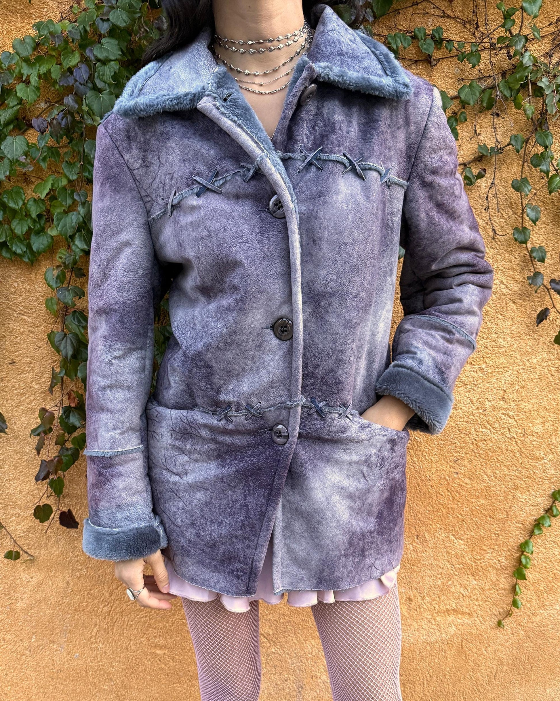 Vintage 2000s faux shearling coat in a rare purple-blue-gray tie-dye abstract finish. Features a soft faux fur collar and cuffs, button closure, side pockets, and decorative criss-cross stitch detailing for a dystopic, cyber-grunge aesthetic. The brushed faux-suede surface gives it a distressed Y2K look, while the straight silhouette makes it easy for winter layering and festival styling. A standout early-2000s piece for streetwear, weird-girl outfits, and bold cold-weather looks.
