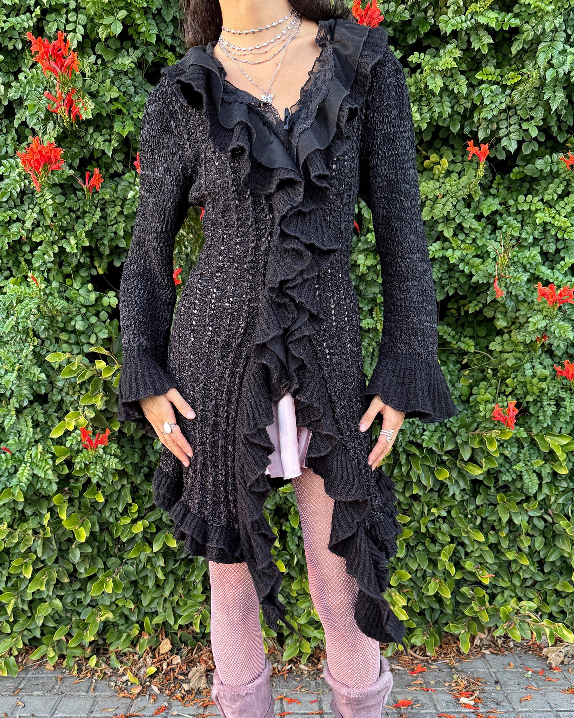 Vintage 2000s black longline cardigan featuring a dramatic ruffle neckline with lace details, bell-sleeve ruffle cuffs and a toggle front closure. The asymmetric pointy hem adds a whimsical, witchy silhouette, while the stretchy cotton–wool–polyamide blend creates a cozy, comfortable fit perfect for winter layering. A stunning fairy-goth statement knit jacket that blends elegance, softness and dark romantic style. Great vintage condition.