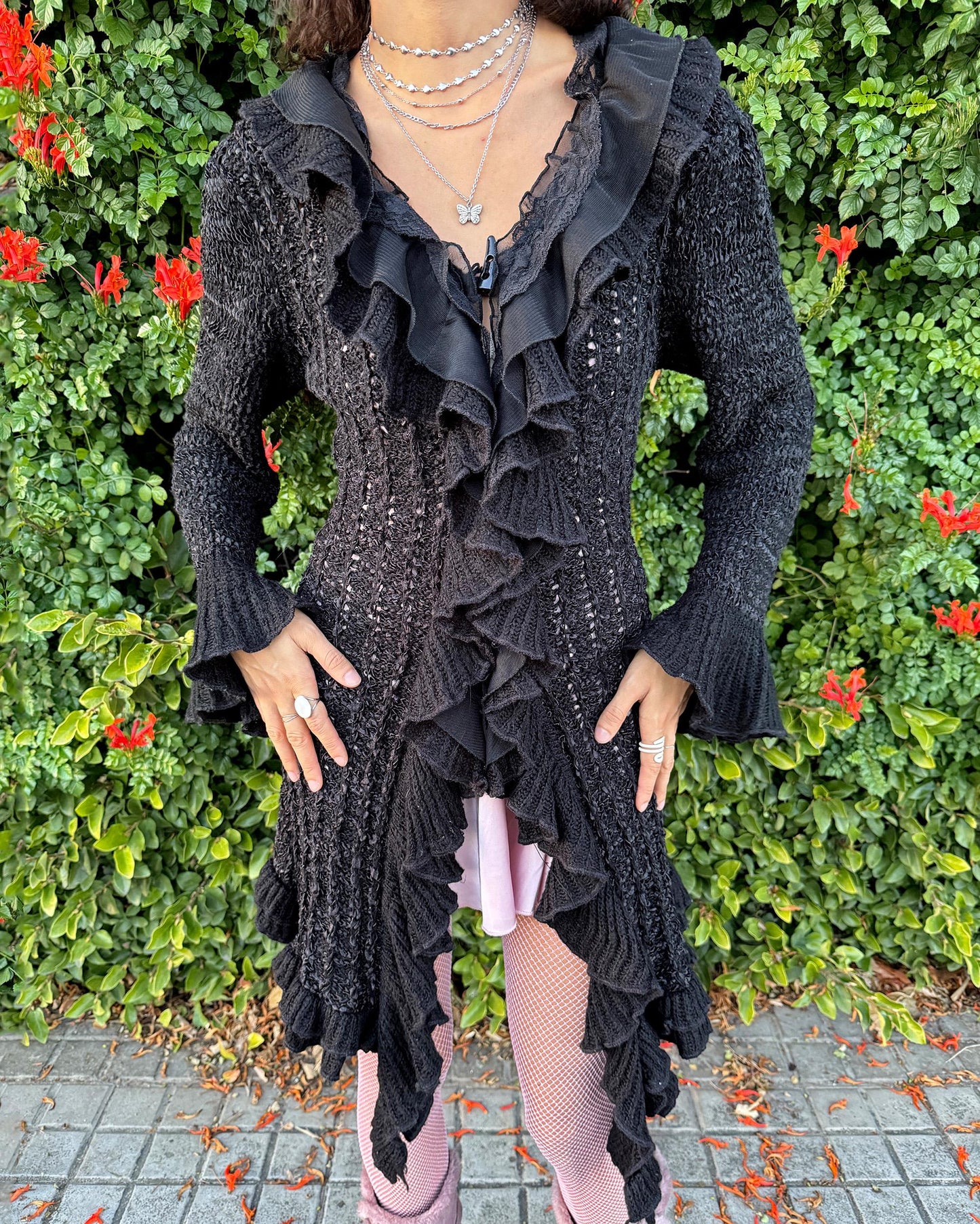 Vintage 2000s black longline cardigan featuring a dramatic ruffle neckline with lace details, bell-sleeve ruffle cuffs and a toggle front closure. The asymmetric pointy hem adds a whimsical, witchy silhouette, while the stretchy cotton–wool–polyamide blend creates a cozy, comfortable fit perfect for winter layering. A stunning fairy-goth statement knit jacket that blends elegance, softness and dark romantic style. Great vintage condition.