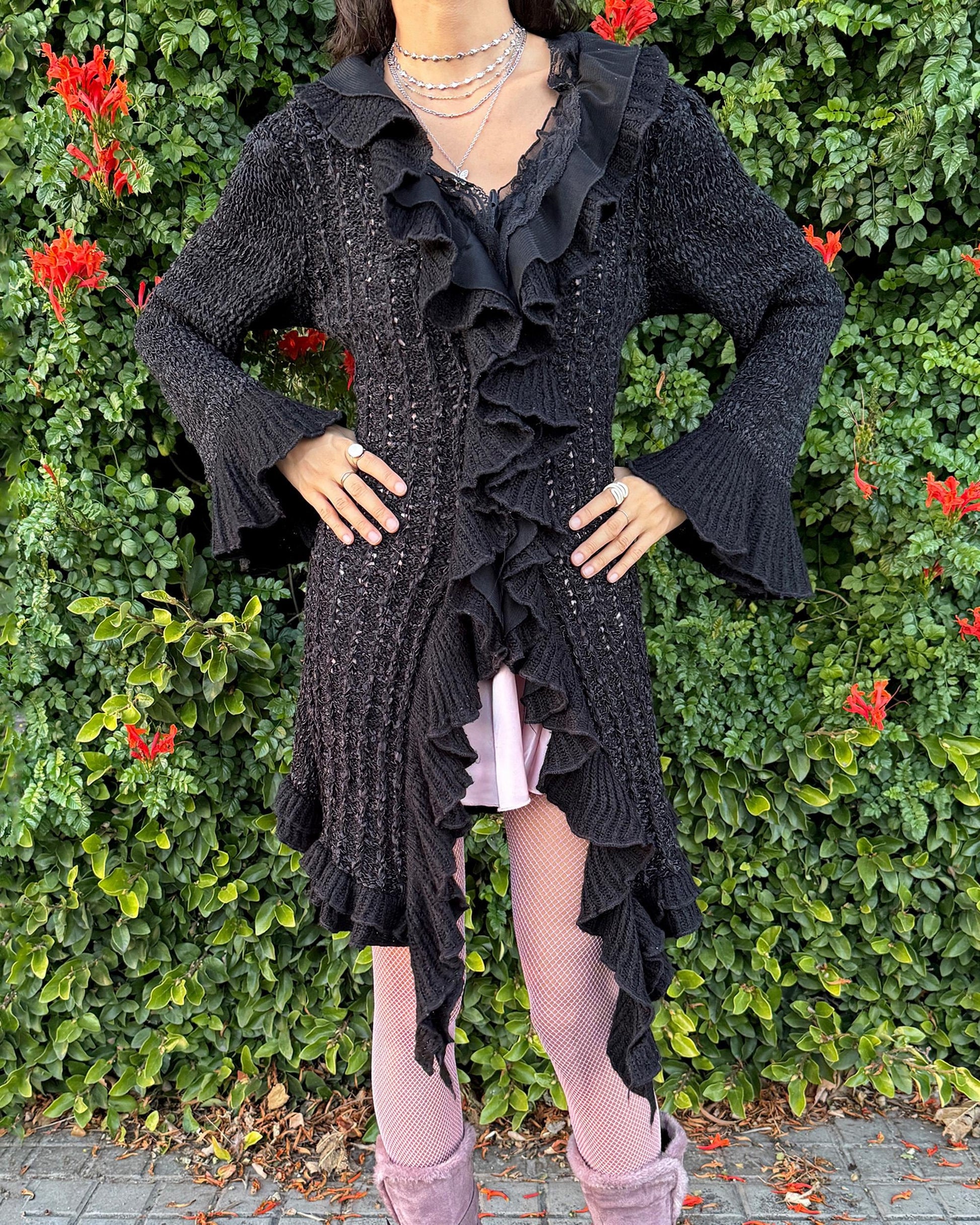 Vintage 2000s black longline cardigan featuring a dramatic ruffle neckline with lace details, bell-sleeve ruffle cuffs and a toggle front closure. The asymmetric pointy hem adds a whimsical, witchy silhouette, while the stretchy cotton–wool–polyamide blend creates a cozy, comfortable fit perfect for winter layering. A stunning fairy-goth statement knit jacket that blends elegance, softness and dark romantic style. Great vintage condition.