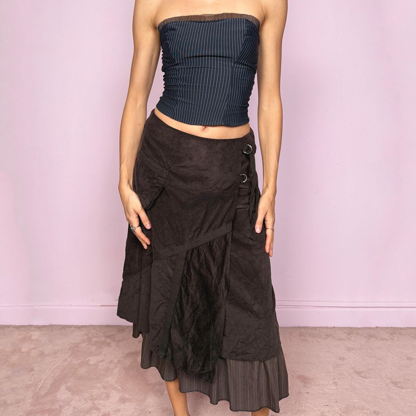 Vintage 2000s dark brown midi skirt with a layered wrap design and ruffled striped hem. Features decorative tie details, a back zipper closure, and a pocket for a deconstructed look. Ideal for earthy fairycore, bohemian, avant-garde, or cyber futuristic styling. Can be worn mid to low rise for size XXS–XS or high waisted for size S. Made from corduroy cotton and polyester. Great vintage condition.