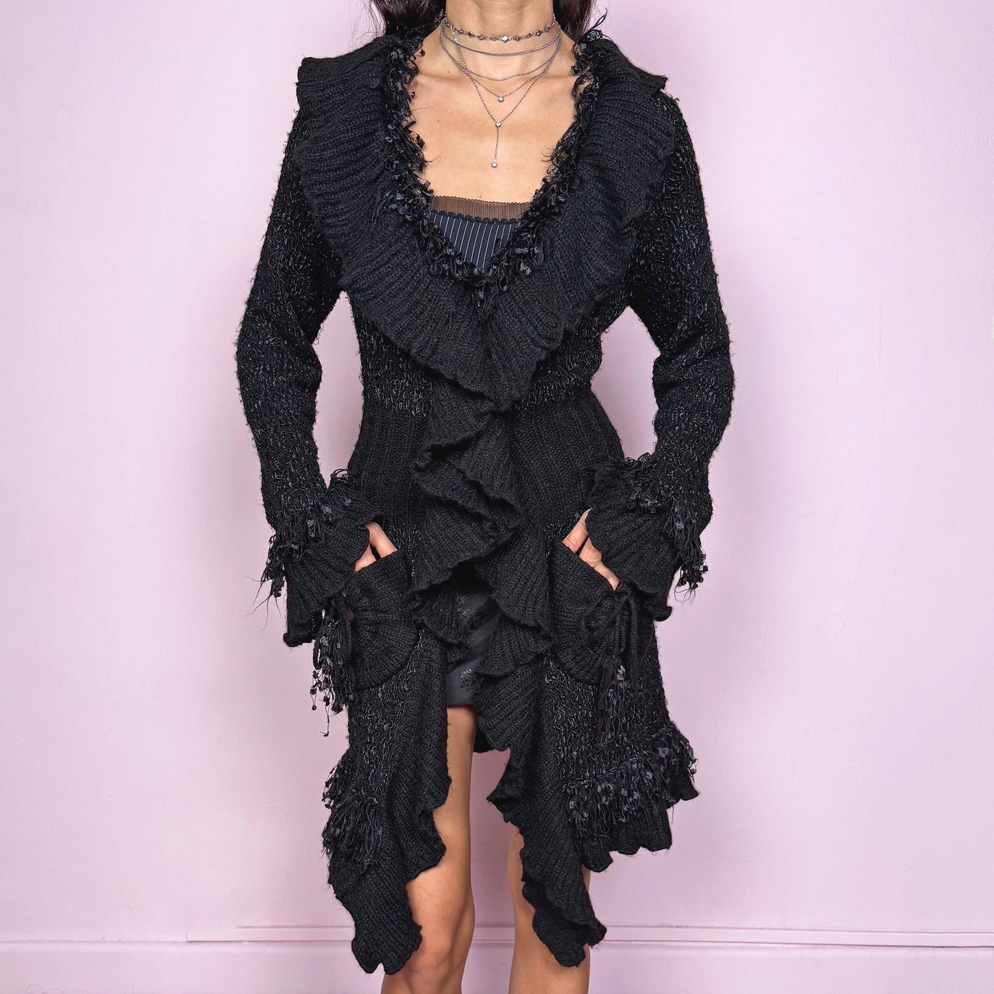 Witchy vintage 2000s black longline cardigan with dramatic ruffle neckline and dark romantic rococo style. Features bell sleeves with ruffle cuffs, two-button front closure, pockets, and asymmetric pointy hem. Whimsical fairycore silhouette, stretchy fabric for cozy winter layering. Made from polyester acrylic blend. Great vintage condition.