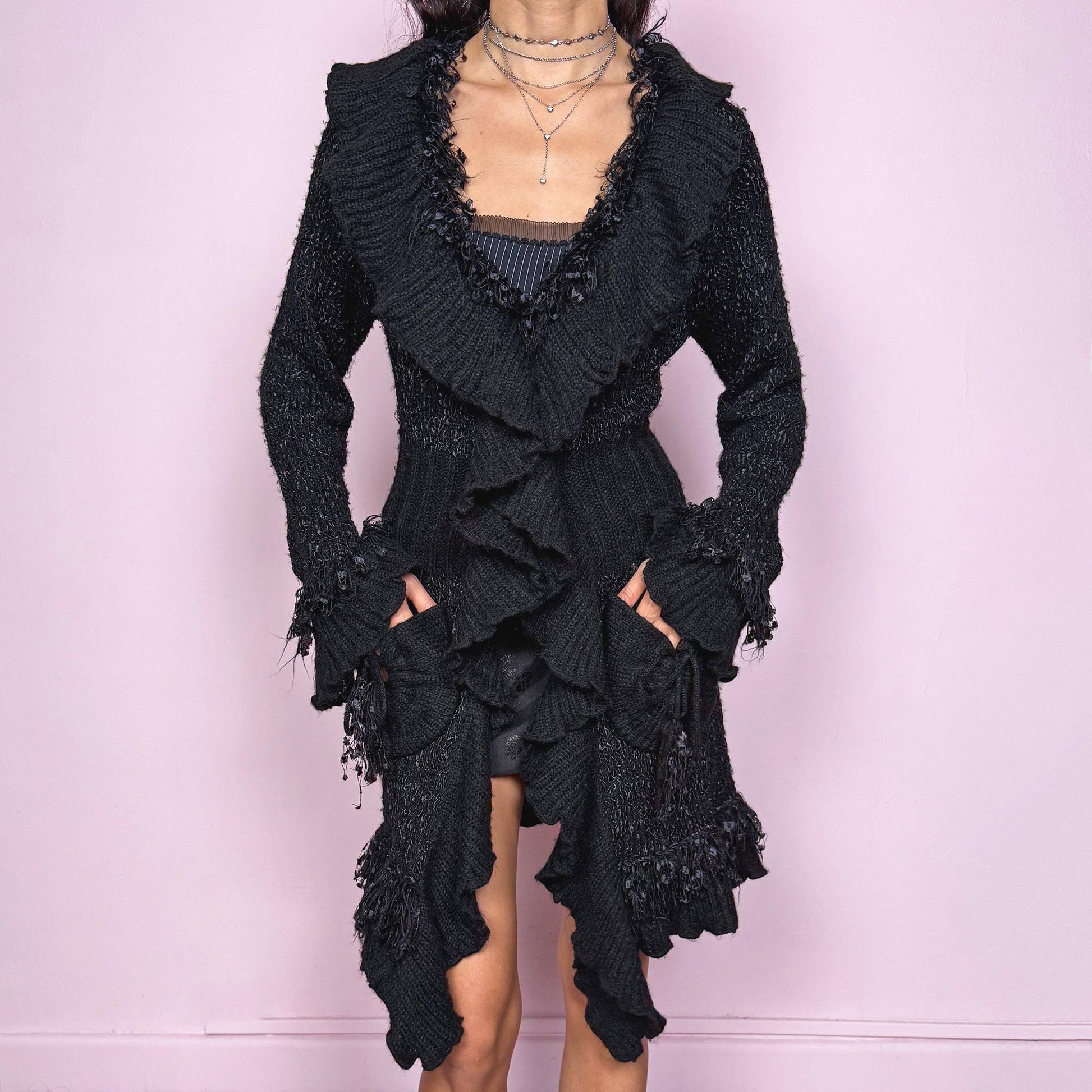 Witchy vintage 2000s black longline cardigan with dramatic ruffle neckline and dark romantic rococo style. Features bell sleeves with ruffle cuffs, two-button front closure, pockets, and asymmetric pointy hem. Whimsical fairycore silhouette, stretchy fabric for cozy winter layering. Made from polyester acrylic blend. Great vintage condition.