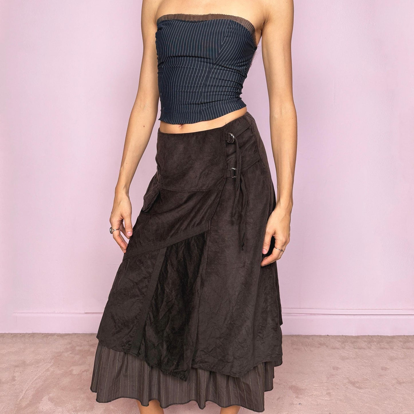Vintage 2000s dark brown midi skirt with a layered wrap design and ruffled striped hem. Features decorative tie details, a back zipper closure, and a pocket for a deconstructed look. Ideal for earthy fairycore, bohemian, avant-garde, or cyber futuristic styling. Can be worn mid to low rise for size XXS–XS or high waisted for size S. Made from corduroy cotton and polyester. Great vintage condition.