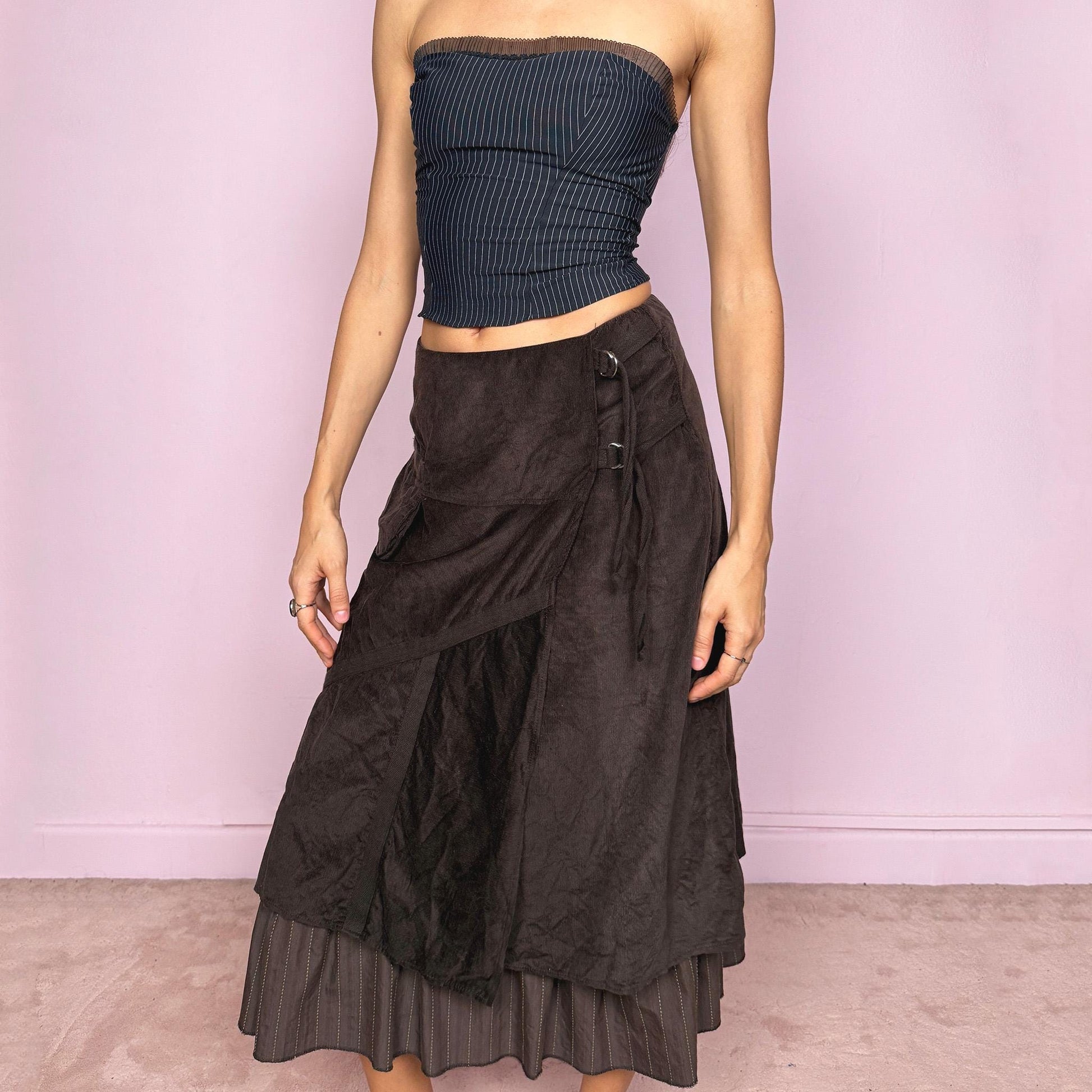 Vintage 2000s dark brown midi skirt with a layered wrap design and ruffled striped hem. Features decorative tie details, a back zipper closure, and a pocket for a deconstructed look. Ideal for earthy fairycore, bohemian, avant-garde, or cyber futuristic styling. Can be worn mid to low rise for size XXS–XS or high waisted for size S. Made from corduroy cotton and polyester. Great vintage condition.