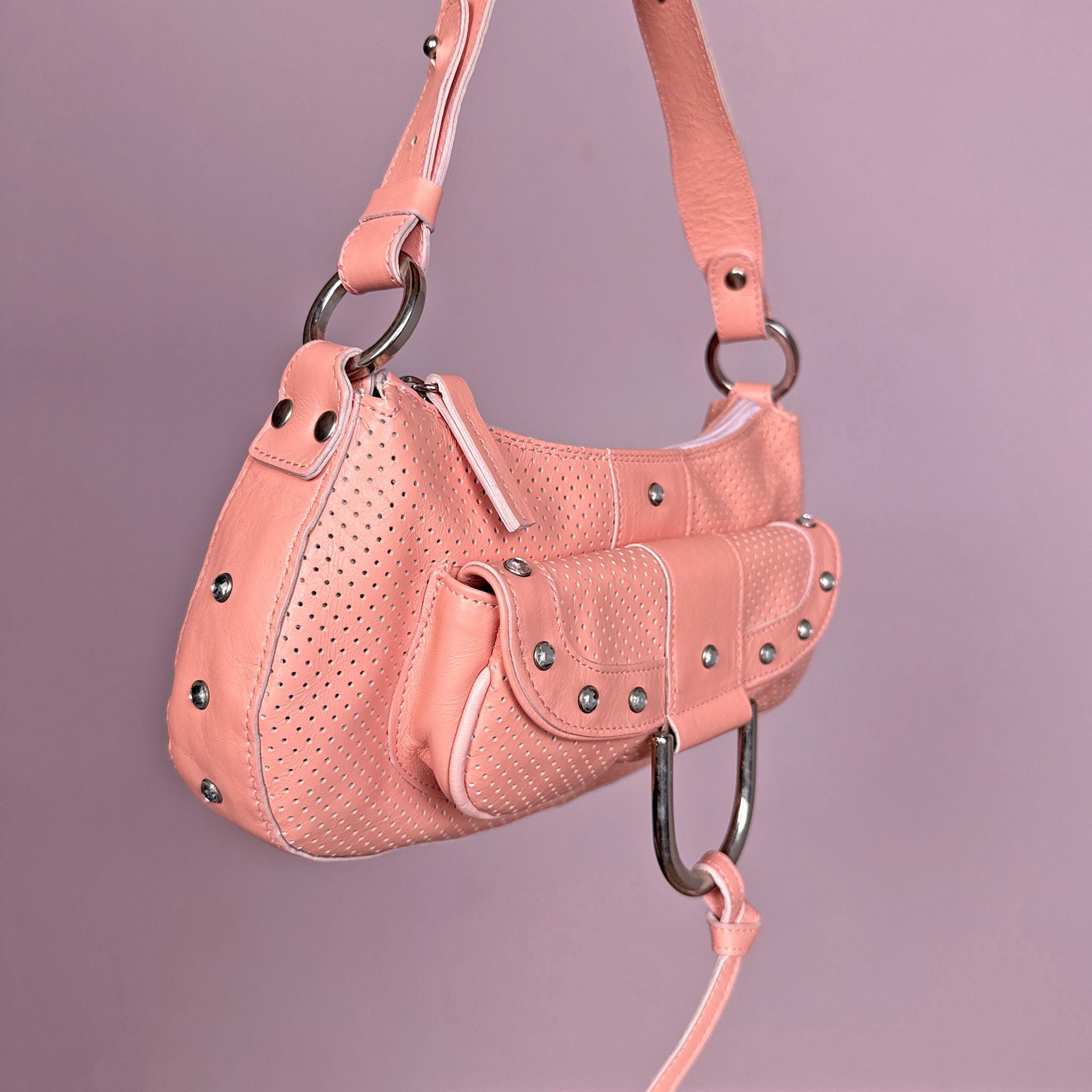 Vintage 2000s pink leather shoulder bag crafted in soft perforated leather with silver tone hardware and a structured baguette silhouette. Features a statement ring detail, front pocket accents with rhinestones, and a zip closure. A feminine yet edgy Y2K party statement piece. Made in Italy. Great vintage condition.