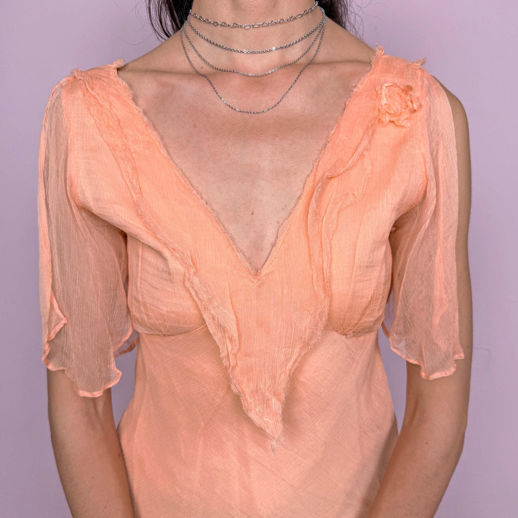 Vintage 2000s pastel light orange-peach fitted top with a deep V-neck, side zip closure, and semi-sheer split flutter sleeves. Romantic, flowy, ethereal design with a soft crinkle texture and a small shoulder rosette, ideal for dreamy summer events or formal looks. Made with a 100% cotton lining and a 100% viscose outer layer. Excellent vintage condition.
