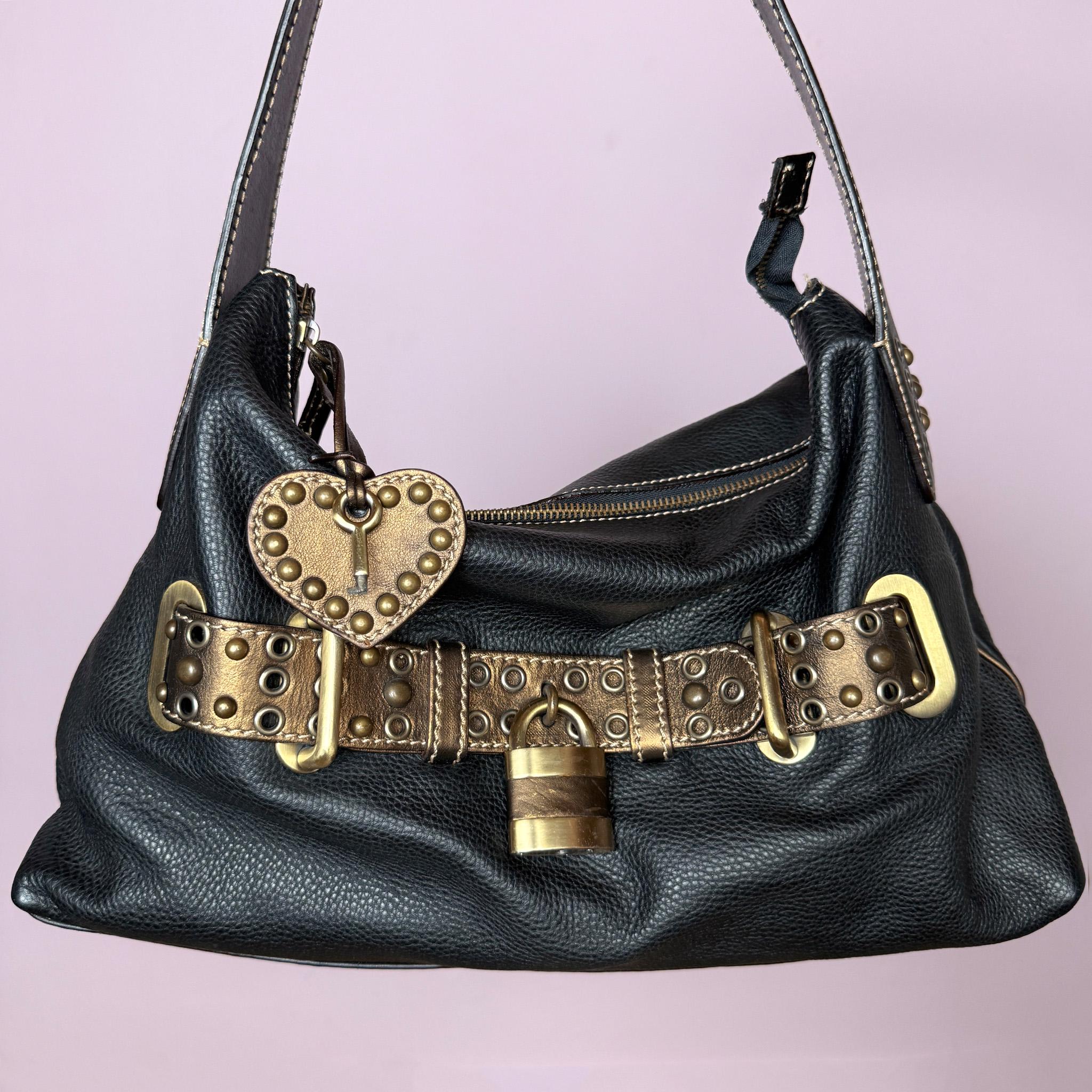 Vintage 2000s black genuine leather shoulder bag made in Italy, Firenze. Features a soft slouchy silhouette, top zip closure, bronze and gold-tone belted front detail, metal eyelets, studded hardware, heart charm, and decorative padlock accent. A unique Y2K Italian leather statement bag with a biker-boho glam and indie grunge feel.