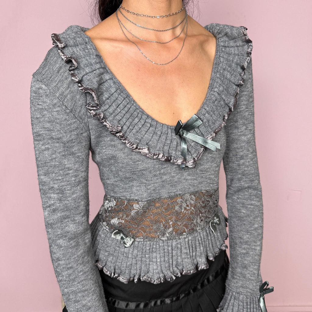 Vintage 2000s gray knit sweater featuring a lace waist panel, delicate ruffle trims, satin bow details, and romantic flared cuffs. Designed with a soft feminine silhouette, deep scoop neckline, and a dreamy coquette and balletcore feel. Made from 70% acrylic and 30% wool. Great vintage condition.