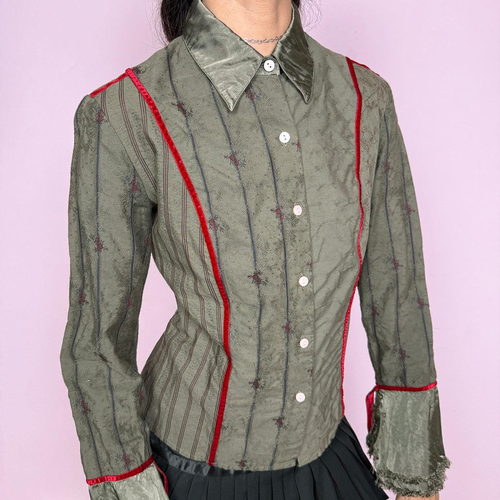 Vintage 2000s muted olive green blouse with subtle floral texture, button-front closure, sharp pointed collar, vertical paneling, contrasting red velvet trim, satin-like collar and cuff details, flared sleeves, and softly frayed edges for elevated earthy styling. Composition label is missing, so the exact fabric is unknown. Great vintage condition.