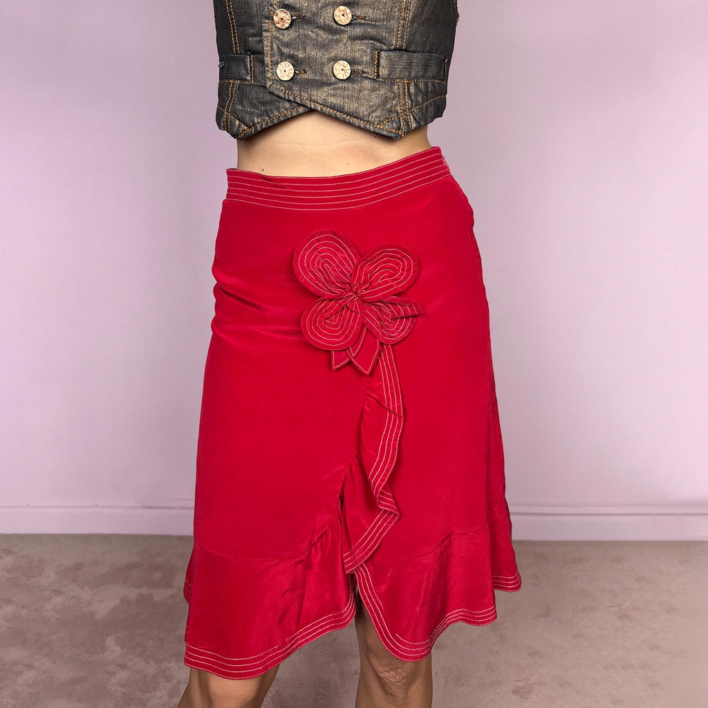 Vintage 2000s Moschino Cheap and Chic red midi skirt with a structured no-stretch fit and side zip closure. It features contrast topstitching through the waistband and hem, a softly flared ruffle hem, and a dimensional flower appliqué at the front with trailing ribbon-like details that create movement and a playful romantic finish. A striking Italian designer piece, ideal for a bold statement feminine outfit. Fits high-waisted for size S. Crafted with a 100% silk
