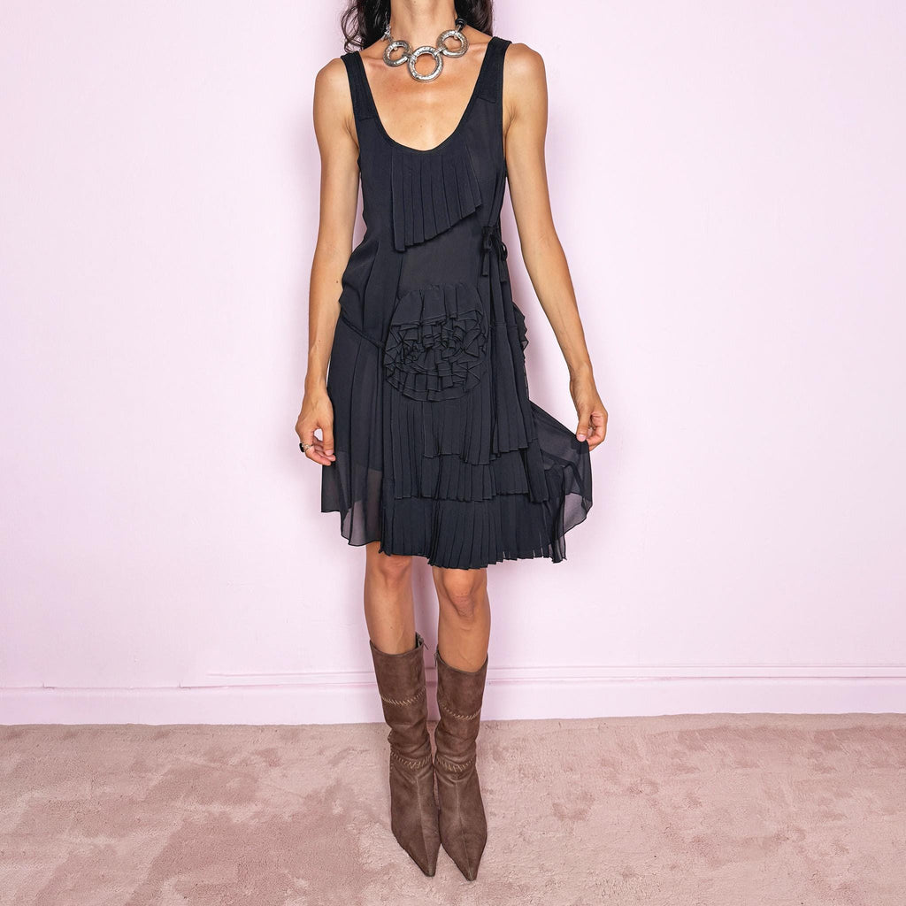 Vintage 2000s black midi dress with a soft sheer layered finish. It features a sleeveless scoop neckline, relaxed shift silhouette, side tie details, and an asymmetric overlay decorated with sculptural pleated appliqués, rosette-style fabric detailing, and layered chiffon panels that add depth and movement. A refined Italian piece with an avant-garde, deconstructed art-to-wear feel, ideal for a minimal elegant evening look. Made by Patrizia Pepe from 100% silk