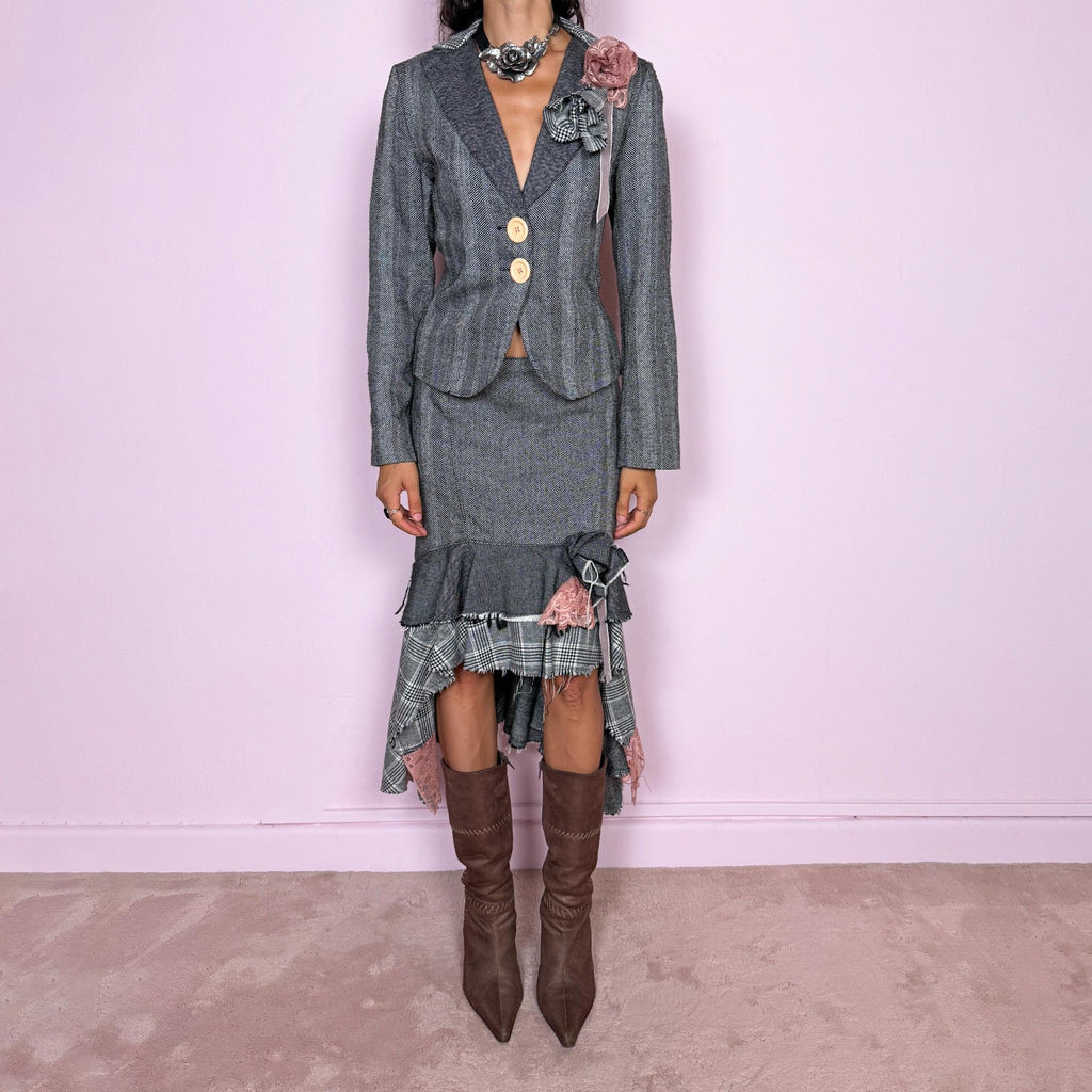 Vintage 2000s two-piece skirt suit crafted from an 80% wool and 20% polyamide blend with no stretch. The fitted gray herringbone blazer features contrasting oversized buttons, wide lapels, and sculptural fabric flower embellishments in dusty pink and monochrome checks, while the matching mid-rise midi skirt combines multiple herringbone and plaid panels with distressed raw edges, asymmetric cascading ruffles, romantic floral appliqué details, and a side zip closure.