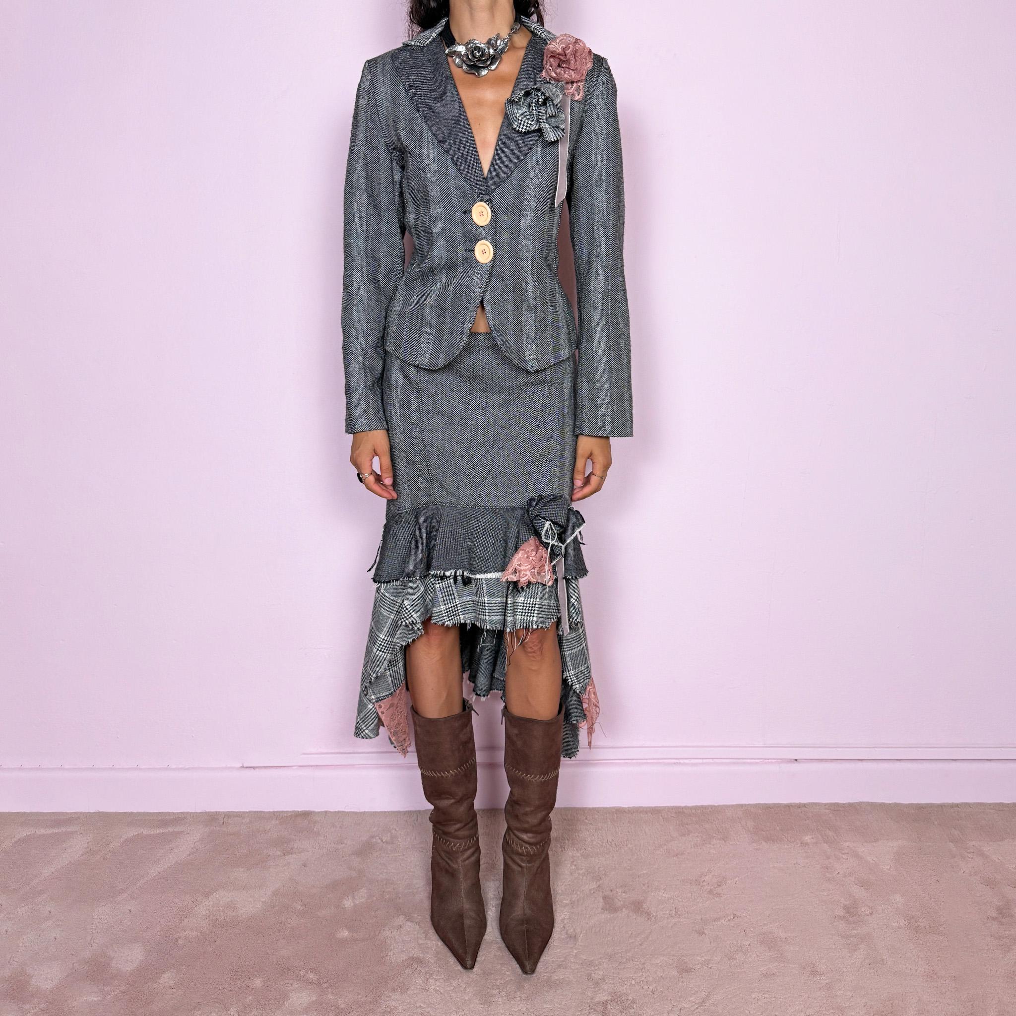 Vintage 2000s two-piece skirt suit crafted from an 80% wool and 20% polyamide blend with no stretch. The fitted gray herringbone blazer features contrasting oversized buttons, wide lapels, and sculptural fabric flower embellishments in dusty pink and monochrome checks, while the matching mid-rise midi skirt combines multiple herringbone and plaid panels with distressed raw edges, asymmetric cascading ruffles, romantic floral appliqué details, and a side zip closure.