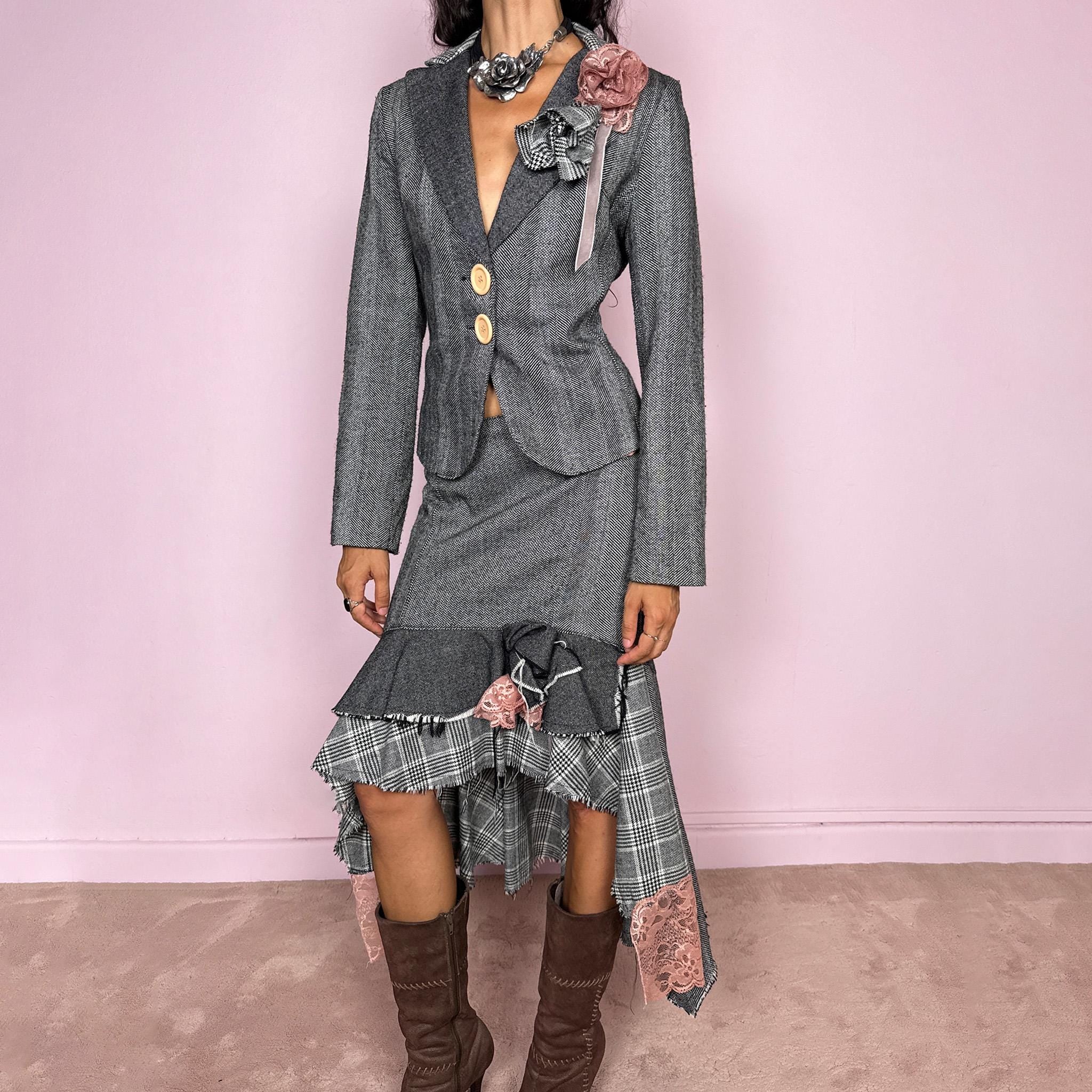 Vintage 2000s two-piece skirt suit crafted from an 80% wool and 20% polyamide blend with no stretch. The fitted gray herringbone blazer features contrasting oversized buttons, wide lapels, and sculptural fabric flower embellishments in dusty pink and monochrome checks, while the matching mid-rise midi skirt combines multiple herringbone and plaid panels with distressed raw edges, asymmetric cascading ruffles, romantic floral appliqué details, and a side zip closure.