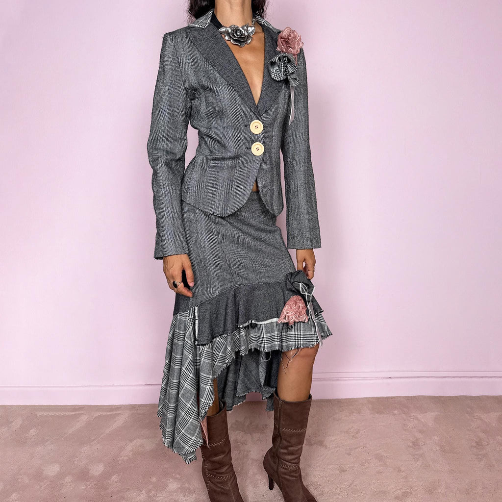 Vintage 2000s two-piece skirt suit crafted from an 80% wool and 20% polyamide blend with no stretch. The fitted gray herringbone blazer features contrasting oversized buttons, wide lapels, and sculptural fabric flower embellishments in dusty pink and monochrome checks, while the matching mid-rise midi skirt combines multiple herringbone and plaid panels with distressed raw edges, asymmetric cascading ruffles, romantic floral appliqué details, and a side zip closure.