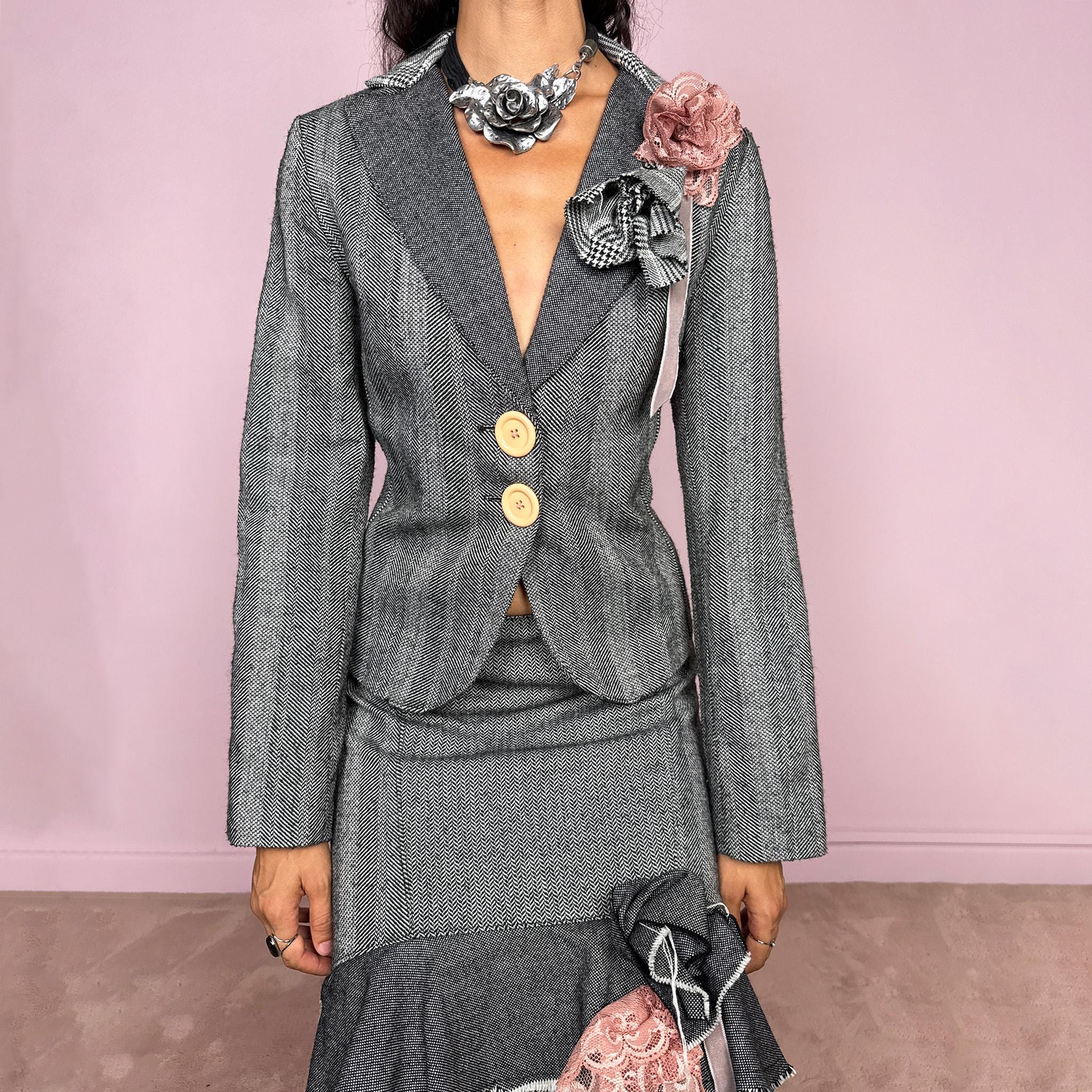 Vintage 2000s two-piece skirt suit crafted from an 80% wool and 20% polyamide blend with no stretch. The fitted gray herringbone blazer features contrasting oversized buttons, wide lapels, and sculptural fabric flower embellishments in dusty pink and monochrome checks, while the matching mid-rise midi skirt combines multiple herringbone and plaid panels with distressed raw edges, asymmetric cascading ruffles, romantic floral appliqué details, and a side zip closure.