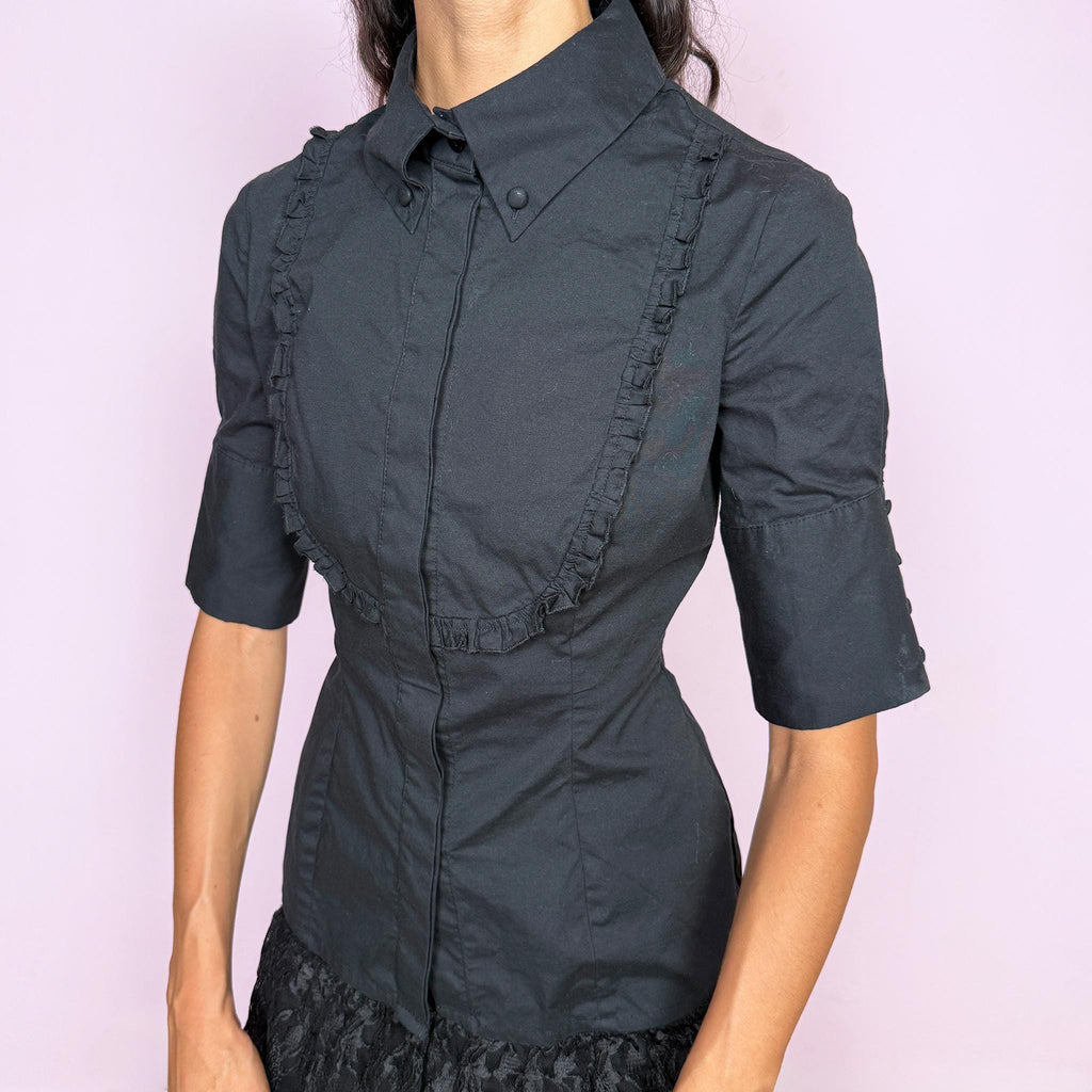Vintage 2000s black fitted shirt top crafted from a slightly stretchy blend of 97% cotton and 3% elastane. Featuring a structured pointed collar, concealed front button closure, and sculpted panel seams that create a sharply tailored silhouette, finished with ruffled bib detailing across the chest and short sleeves. A refined piece balancing formal tailoring with subtle romantic detailing, ideal for elegant minimalist or dark feminine styling. By Spanish designer Adolfo Domínguez.