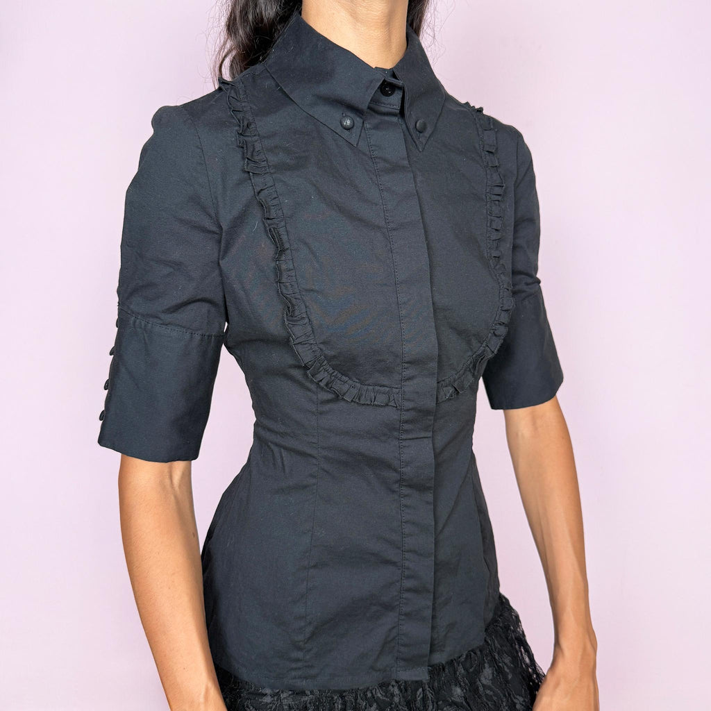 Vintage 2000s black fitted shirt top crafted from a slightly stretchy blend of 97% cotton and 3% elastane. Featuring a structured pointed collar, concealed front button closure, and sculpted panel seams that create a sharply tailored silhouette, finished with ruffled bib detailing across the chest and short sleeves. A refined piece balancing formal tailoring with subtle romantic detailing, ideal for elegant minimalist or dark feminine styling. By Spanish designer Adolfo Domínguez.