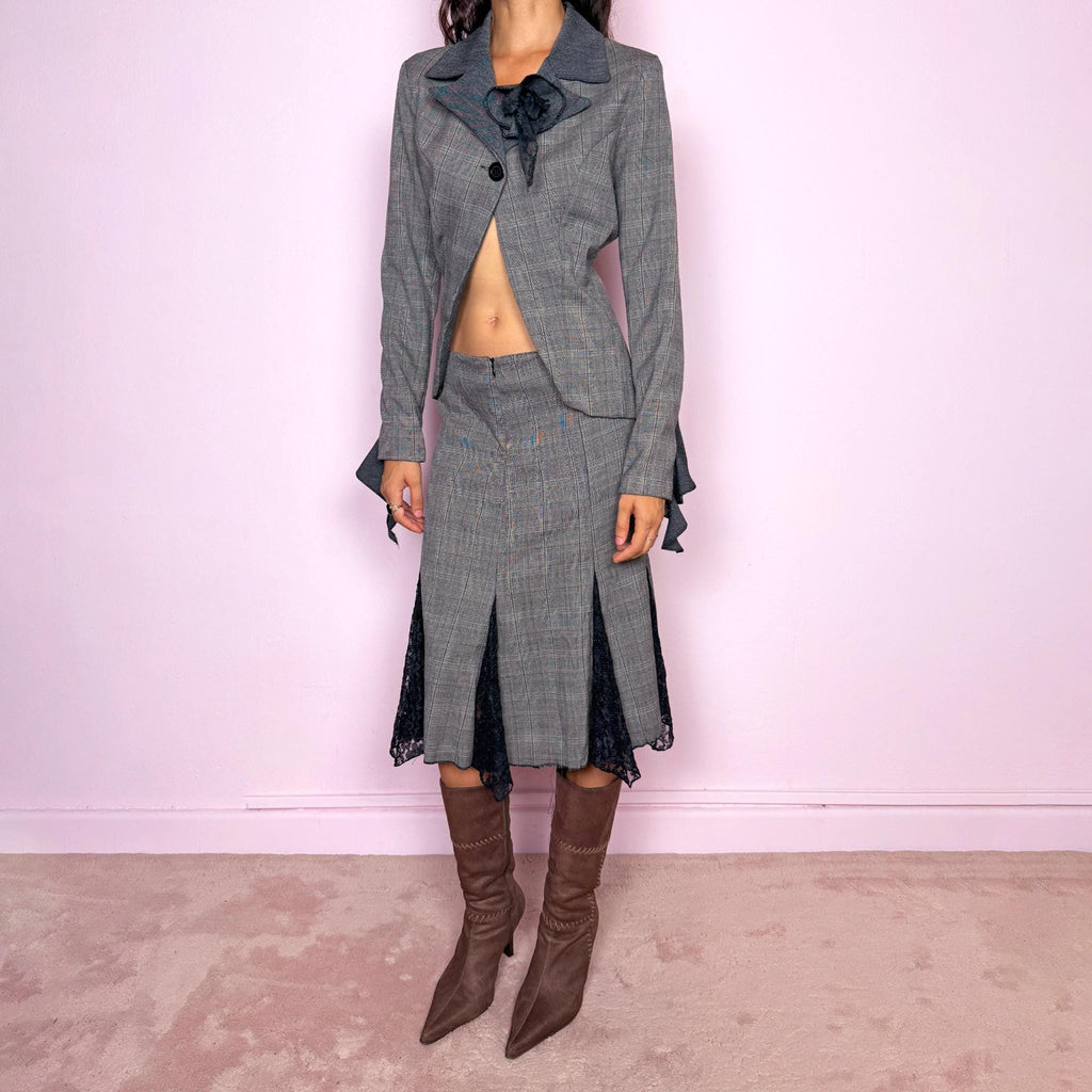 Vintage 2000s gray two-piece skirt suit. The fitted blazer is crafted from a 50% wool and 50% viscose blend and features contrasting darker lapels, a single-button closure, sculptural floral appliqué, and an asymmetric cutaway front. The matching midi skirt is crafted from a slightly stretchy polyester-viscose blend and features a front zip closure, mid-rise fit, and dramatic black lace godet-style inserts through the lower panels, creating a deconstructed tailored silhouette.