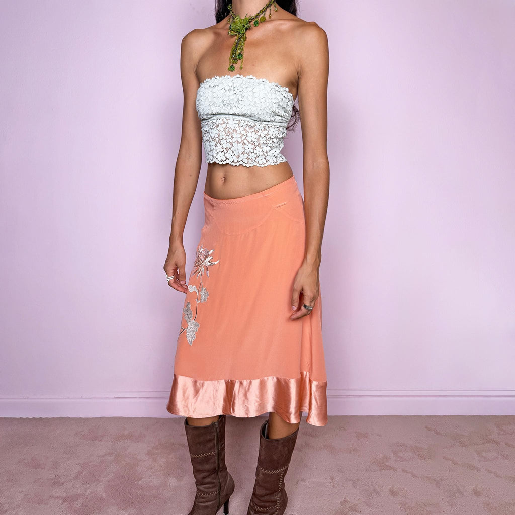Vintage 2000s pastel salmon-orange midi skirt crafted from a fluid, non-stretch fabric with a silk-like feel, featuring delicate white floral embroidery, a flared silhouette, satin trim at the hem, and a satin lining. The soft salmon tone and botanical embroidery give it a dreamy, romantic bohemian aesthetic. The composition label is missing, so the exact fabric is unknown, though it has the look and feel of silk. From a Danish brand.