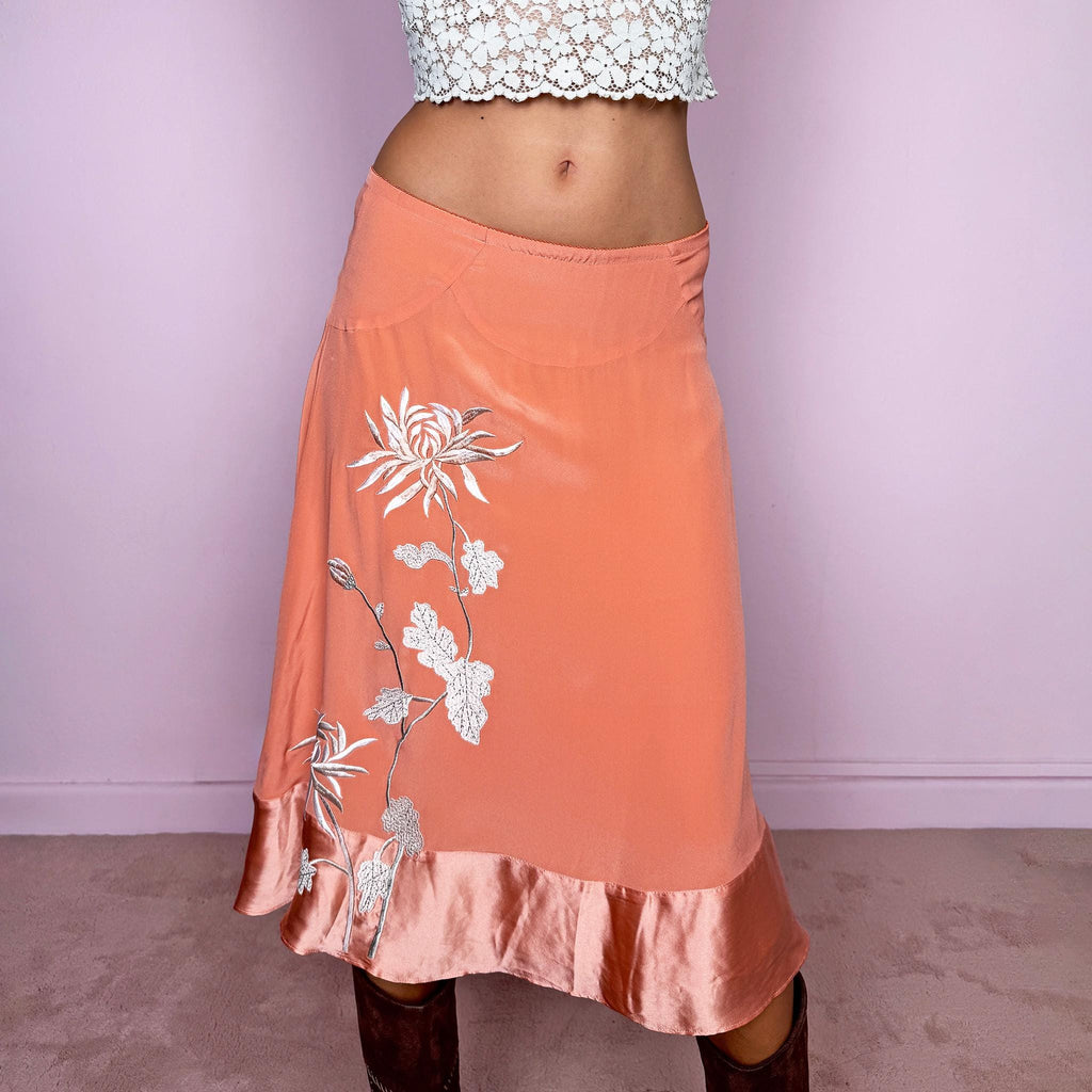 Vintage 2000s pastel salmon-orange midi skirt crafted from a fluid, non-stretch fabric with a silk-like feel, featuring delicate white floral embroidery, a flared silhouette, satin trim at the hem, and a satin lining. The soft salmon tone and botanical embroidery give it a dreamy, romantic bohemian aesthetic. The composition label is missing, so the exact fabric is unknown, though it has the look and feel of silk. From a Danish brand.