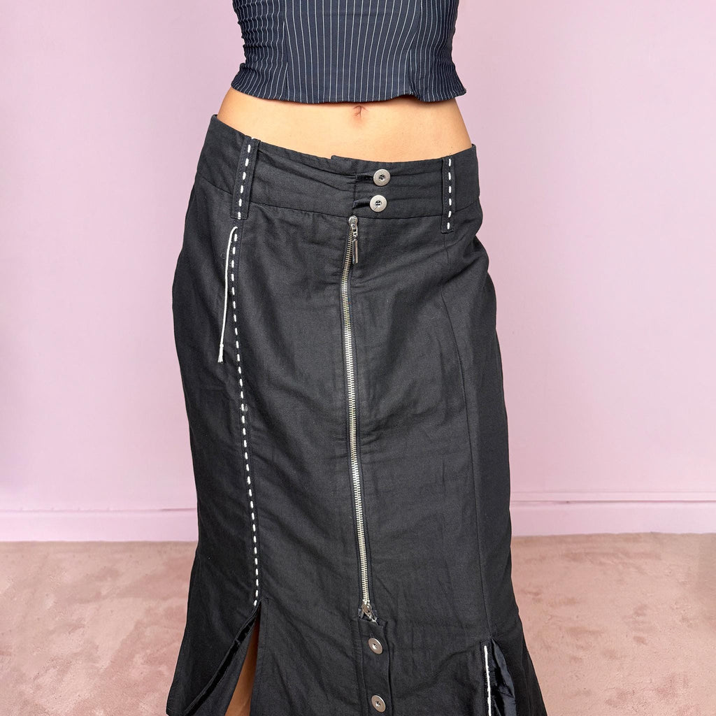 Vintage 2000s black midi skirt featuring an exposed full-length front zip, double-button waistband, and contrast white stitching throughout. The lower section opens into sculptural deconstructed panels with additional button and zip detailing, creating an asymmetric, utilitarian avant-garde feel. Can be worn high-waisted for size M or slightly lower on the waist for sizes S and XS. Non-stretch. Made in Holland by a Dutch brand from a structured blend of 52% cotton and 48% linen