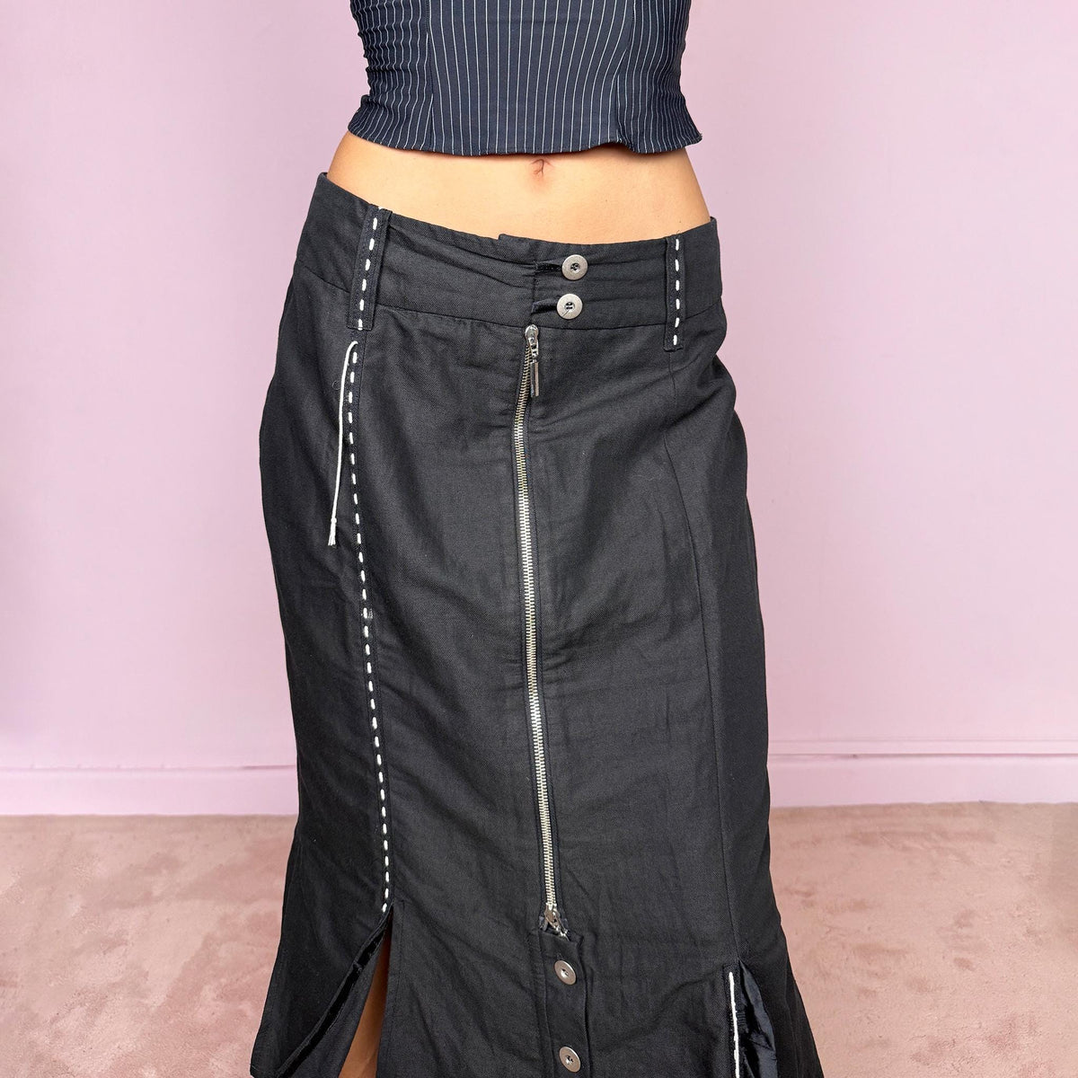 Vintage 2000s black midi skirt featuring an exposed full-length front zip, double-button waistband, and contrast white stitching throughout. The lower section opens into sculptural deconstructed panels with additional button and zip detailing, creating an asymmetric, utilitarian avant-garde feel. Can be worn high-waisted for size M or slightly lower on the waist for sizes S and XS. Non-stretch. Made in Holland by a Dutch brand from a structured blend of 52% cotton and 48% linen