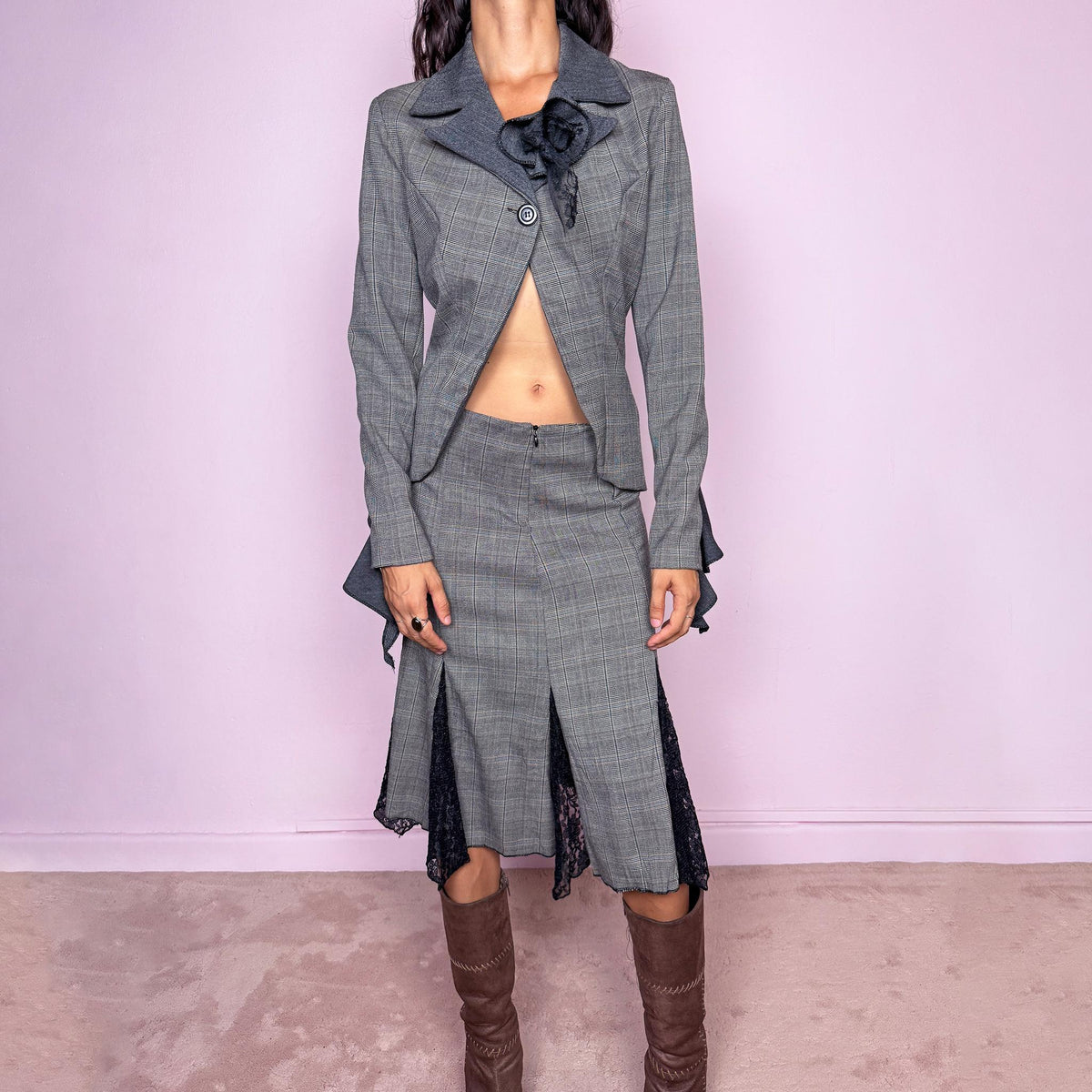 Vintage 2000s gray two-piece skirt suit. The fitted blazer is crafted from a 50% wool and 50% viscose blend and features contrasting darker lapels, a single-button closure, sculptural floral appliqué, and an asymmetric cutaway front. The matching midi skirt is crafted from a slightly stretchy polyester-viscose blend and features a front zip closure, mid-rise fit, and dramatic black lace godet-style inserts through the lower panels, creating a deconstructed tailored silhouette.