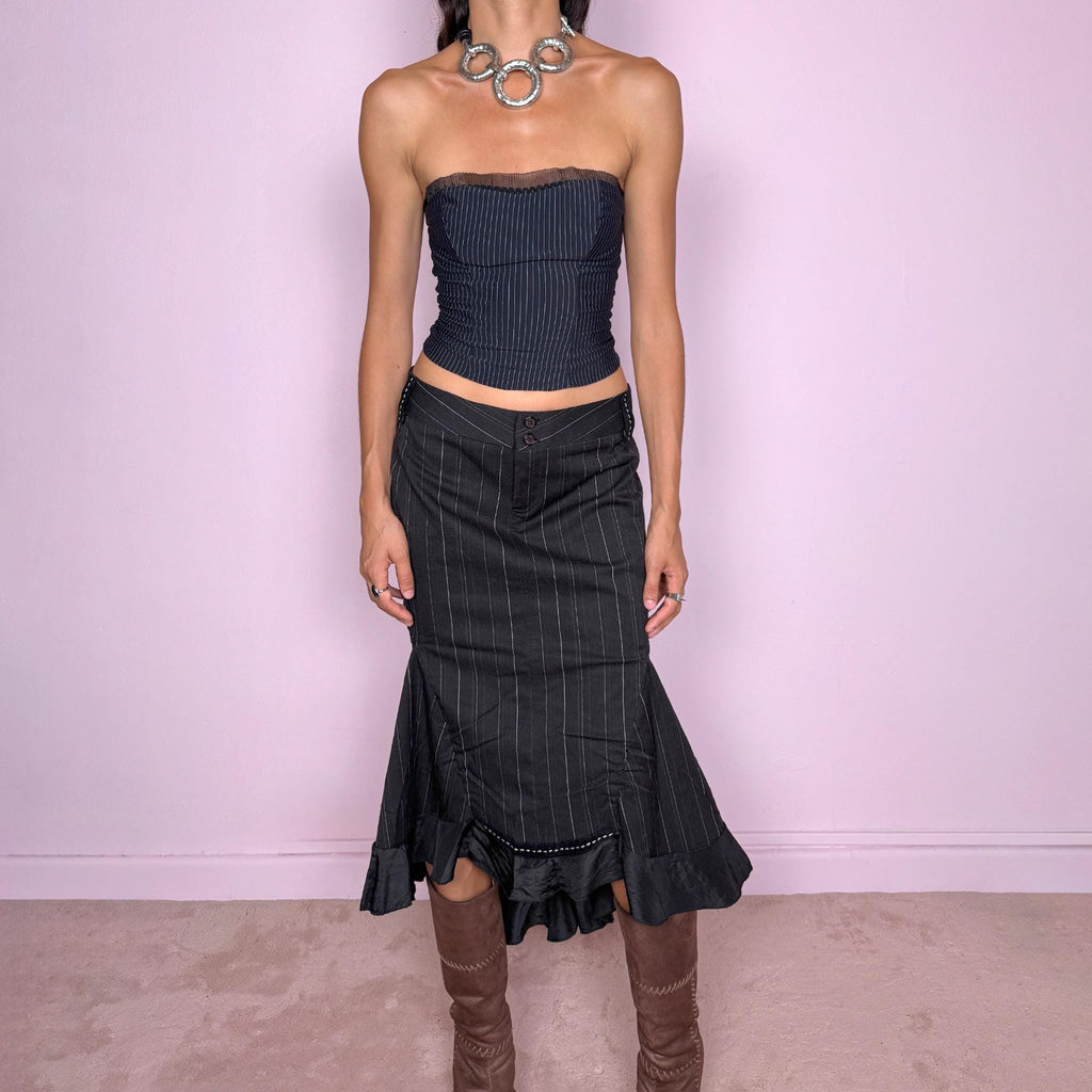 Vintage 2000s black pinstripe midi skirt featuring a fitted low-rise silhouette with a diagonal waistband construction, front zip closure, vertical pinstripes, and sculptural paneling through the lower skirt. Finished with an asymmetric flared hem and contrasting satin-like ruffle trim for a dramatic, deconstructed Y2K silhouette. Made in Italy by an Italian brand from a slightly stretchy blend of 62% polyester, 33% viscose, and 5% elastane. Excellent vintage condition.