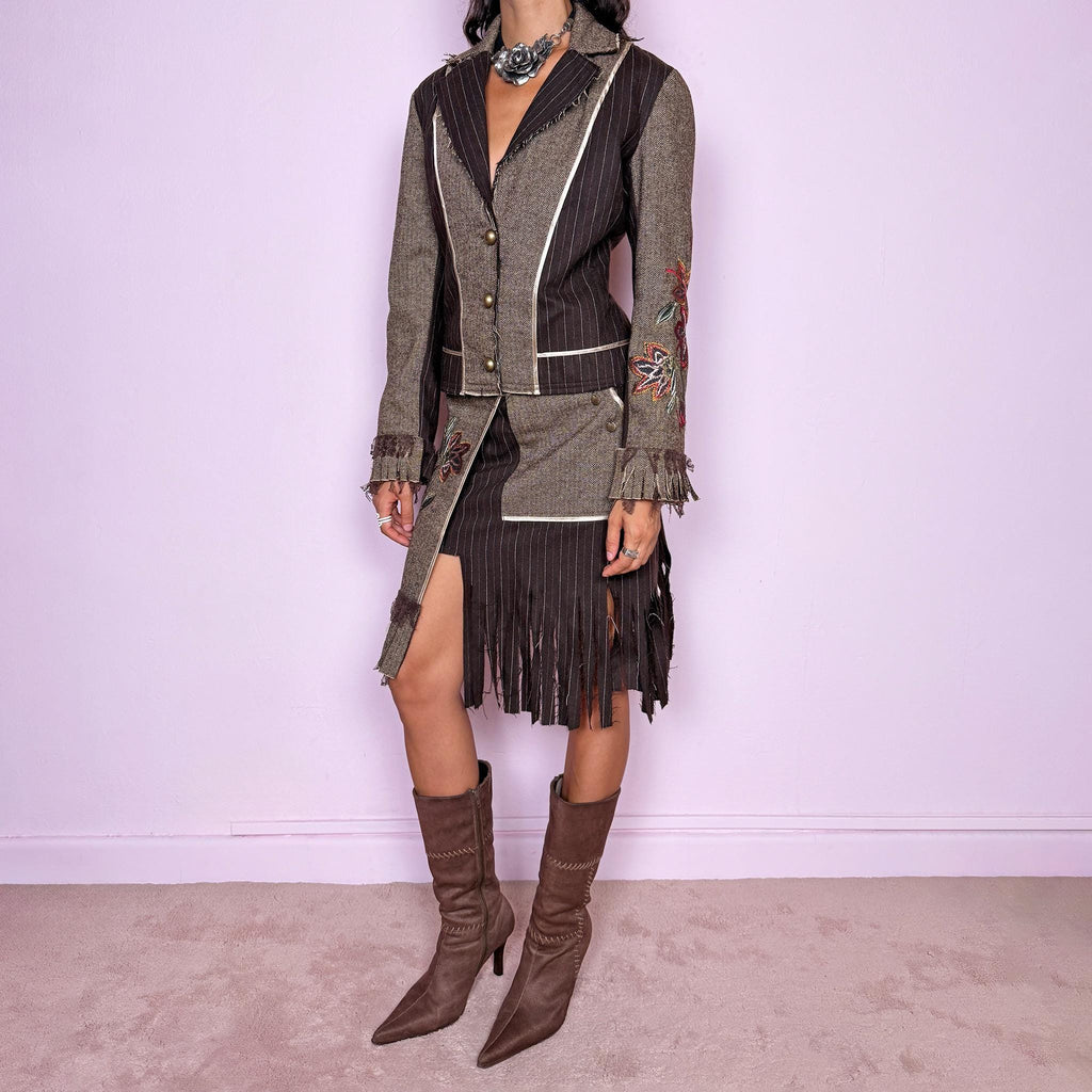 Vintage 2000s deconstructed two-piece skirt suit. The fitted jacket features mixed pinstripe and herringbone panels, raw-edge detailing, contrast piping, floral embroidery, decorative metal buttons, and pleated fringe cuffs. Paired with a matching asymmetric midi skirt featuring layered panel construction, a side zip closure, embroidered floral motifs, and dramatic cut fringe through the lower section. Non-stretch.