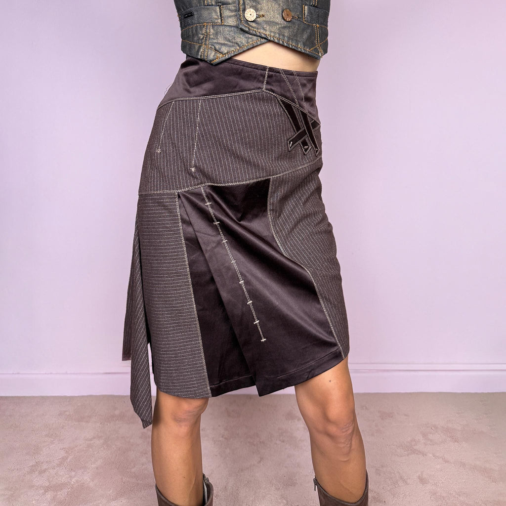 Vintage 2000s brown asymmetric midi skirt featuring a high-waisted, fitted silhouette with contrasting pinstripe and satin-like panels, sculptural draping along one side, and an elongated asymmetric hanging panel. Designed with contrast topstitching, decorative strap detailing, and a side zip closure. Non-stretch. The mix of tailored pinstripes, fluid draping, and deconstructed paneling gives it an avant-garde aesthetic. From a French brand.