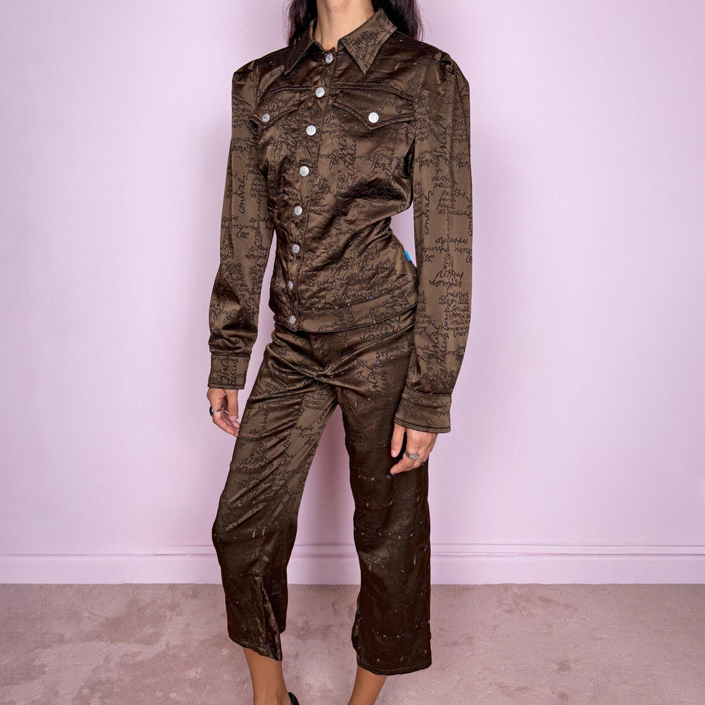 Vintage late 1990s two-piece set from French fashion house Christian Lacroix, from the Jeans de Christian Lacroix line, made in Italy. The set features a brown printed jacket with a glossy, satin-like finish, handwritten-style graphic motif, pointed collar, flap chest pockets, and branded snap-button closure, paired with matching high-waisted cropped trousers with a slim fit through the leg and flared hems. Crafted from a stretchy blend of acetate, polyamide, polyester, and elastane.