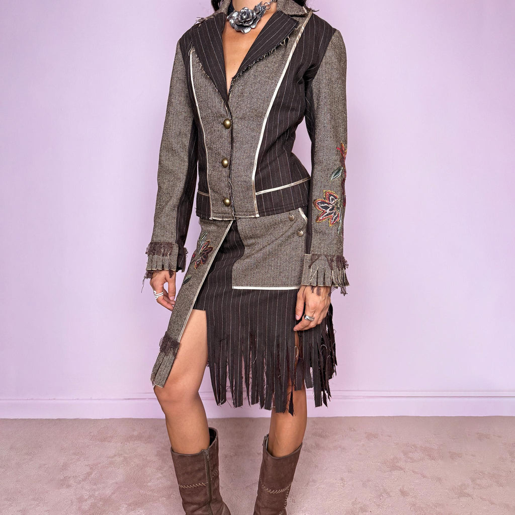 Vintage 2000s deconstructed two-piece skirt suit. The fitted jacket features mixed pinstripe and herringbone panels, raw-edge detailing, contrast piping, floral embroidery, decorative metal buttons, and pleated fringe cuffs. Paired with a matching asymmetric midi skirt featuring layered panel construction, a side zip closure, embroidered floral motifs, and dramatic cut fringe through the lower section. Non-stretch.
