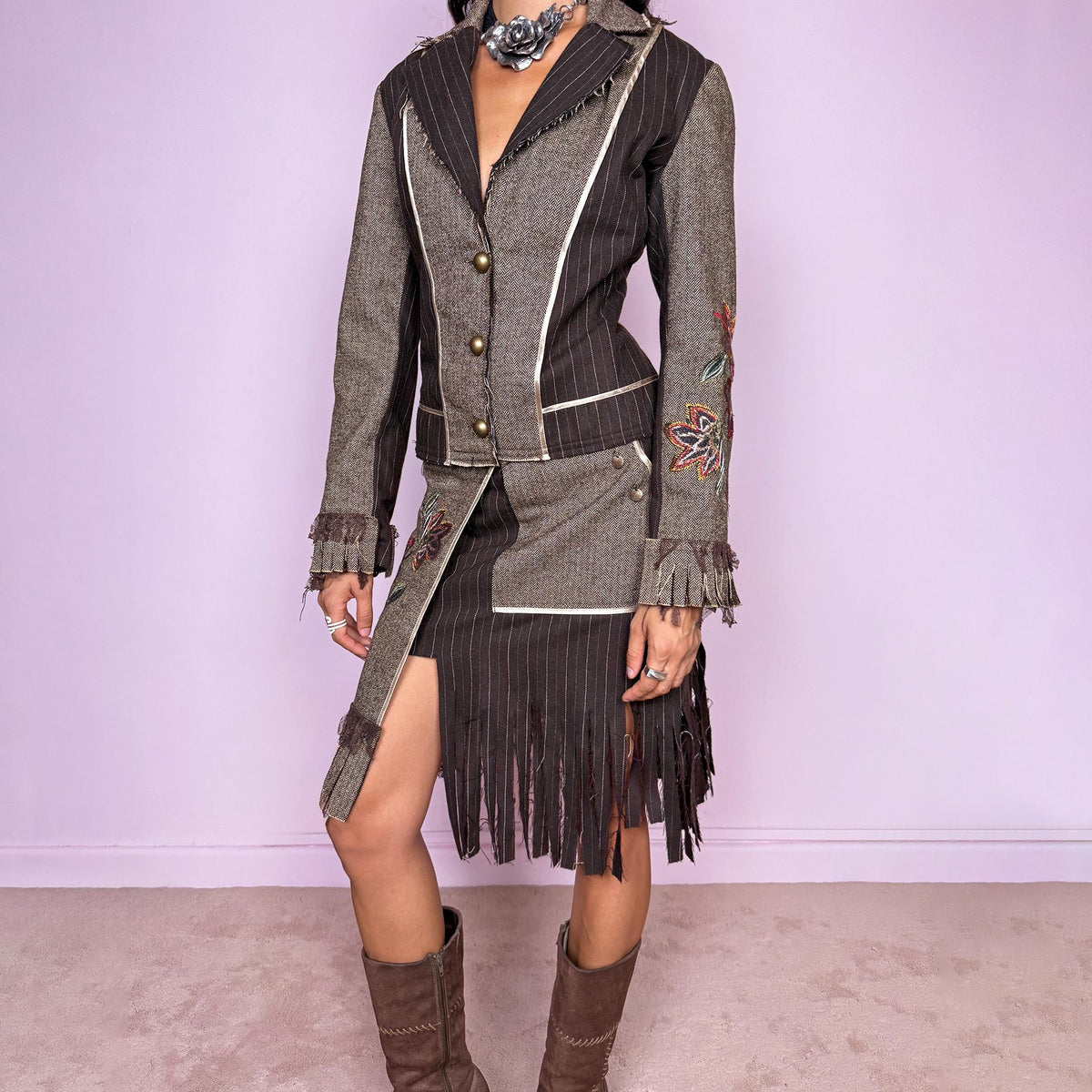 Vintage 2000s deconstructed two-piece skirt suit. The fitted jacket features mixed pinstripe and herringbone panels, raw-edge detailing, contrast piping, floral embroidery, decorative metal buttons, and pleated fringe cuffs. Paired with a matching asymmetric midi skirt featuring layered panel construction, a side zip closure, embroidered floral motifs, and dramatic cut fringe through the lower section. Non-stretch.