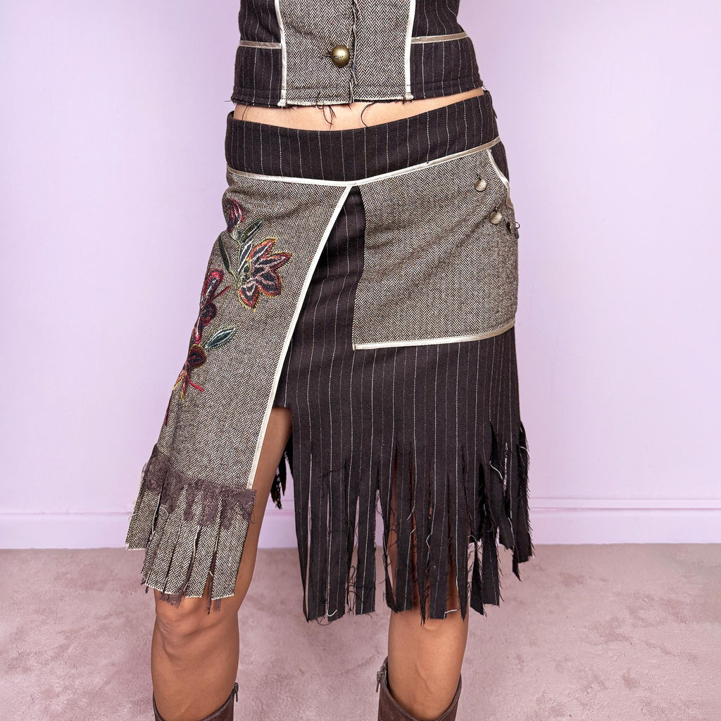 Vintage 2000s deconstructed two-piece skirt suit. The fitted jacket features mixed pinstripe and herringbone panels, raw-edge detailing, contrast piping, floral embroidery, decorative metal buttons, and pleated fringe cuffs. Paired with a matching asymmetric midi skirt featuring layered panel construction, a side zip closure, embroidered floral motifs, and dramatic cut fringe through the lower section. Non-stretch.