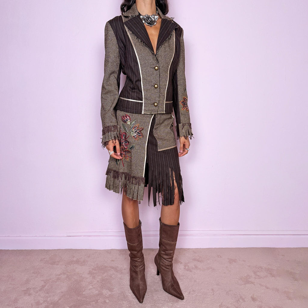 Vintage 2000s deconstructed two-piece skirt suit. The fitted jacket features mixed pinstripe and herringbone panels, raw-edge detailing, contrast piping, floral embroidery, decorative metal buttons, and pleated fringe cuffs. Paired with a matching asymmetric midi skirt featuring layered panel construction, a side zip closure, embroidered floral motifs, and dramatic cut fringe through the lower section. Non-stretch.