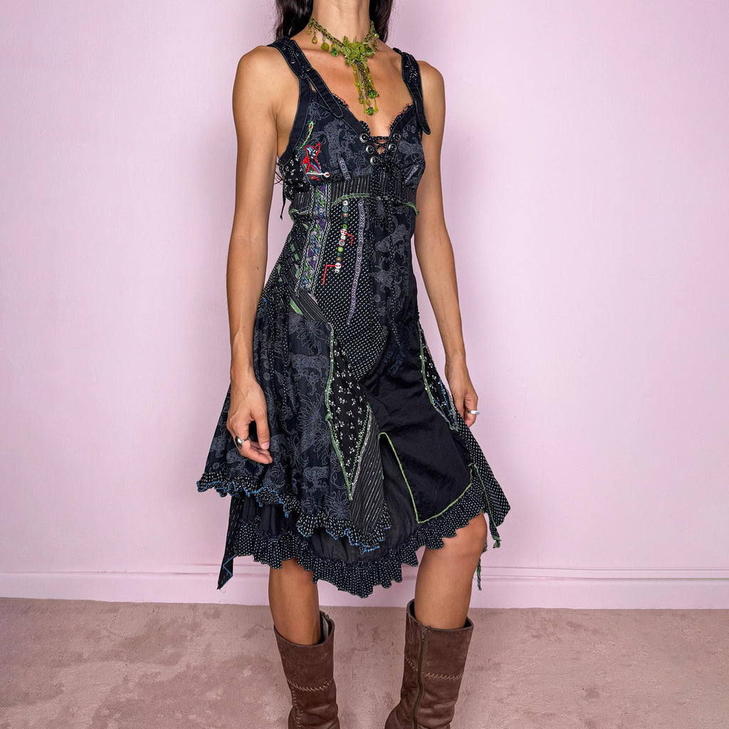 Vintage 2000s black midi dress featuring a deconstructed patchwork construction with mixed pinstripe, floral, and textured panels. Designed with a deep lace-up neckline, asymmetric layered skirt, raw-edge detailing, decorative stitching, and a dramatic draped side panel. Finished with a side zip closure. Non-stretch. The irregular construction and mixed textures are ideal for ragged-doll, avant-garde looks.