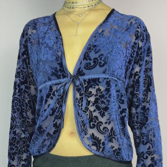 The Vintage 90s Blue Velvet Sheer Blouse is a romantic sheer velvet top featuring a flocked baroque floral pattern on deep navy blue. Designed with a plunging V-neckline, long sleeves, and a delicate tie-front closure, this blouse is perfect for night-out looks.