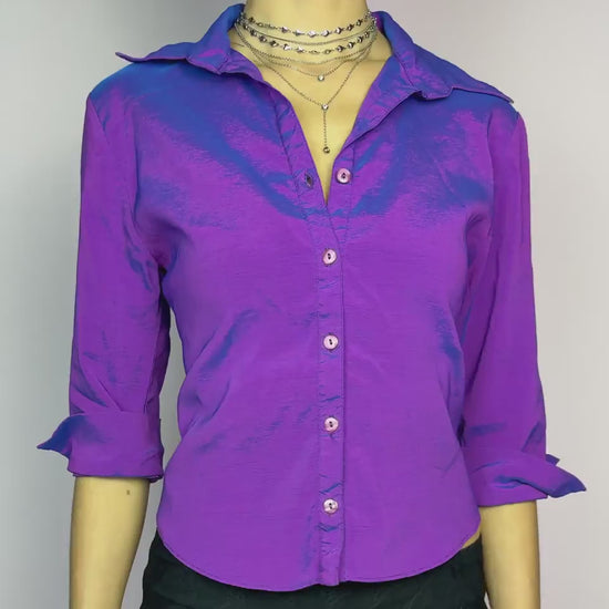 The Y2K Purple Iridescent Blouse is a vintage 2000s button-up top featuring a shimmery, iridescent stretchy fabric that shifts between purple and blue tones depending on the light. Designed with a structured collar, a fitted silhouette, and rolled-up 3/4 sleeves for a casual-chic look. Perfect for night outs and parties.