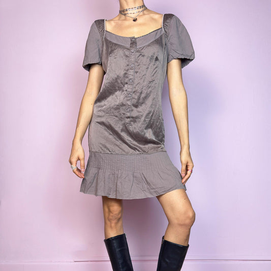 This vintage 2000s mini dress comes in a soft taupe (brown-grey) cotton and silk blend with a satin-like sheen. It features puff sleeves, a square neckline, and buttons down the front. The dropped waist flows into a pleated mini hem for a flirty silhouette. Lightweight and breathable, ideal for warm-weather dressing. A romantic piece with coquette undertones, blending softness and structure in a quietly nostalgic way.