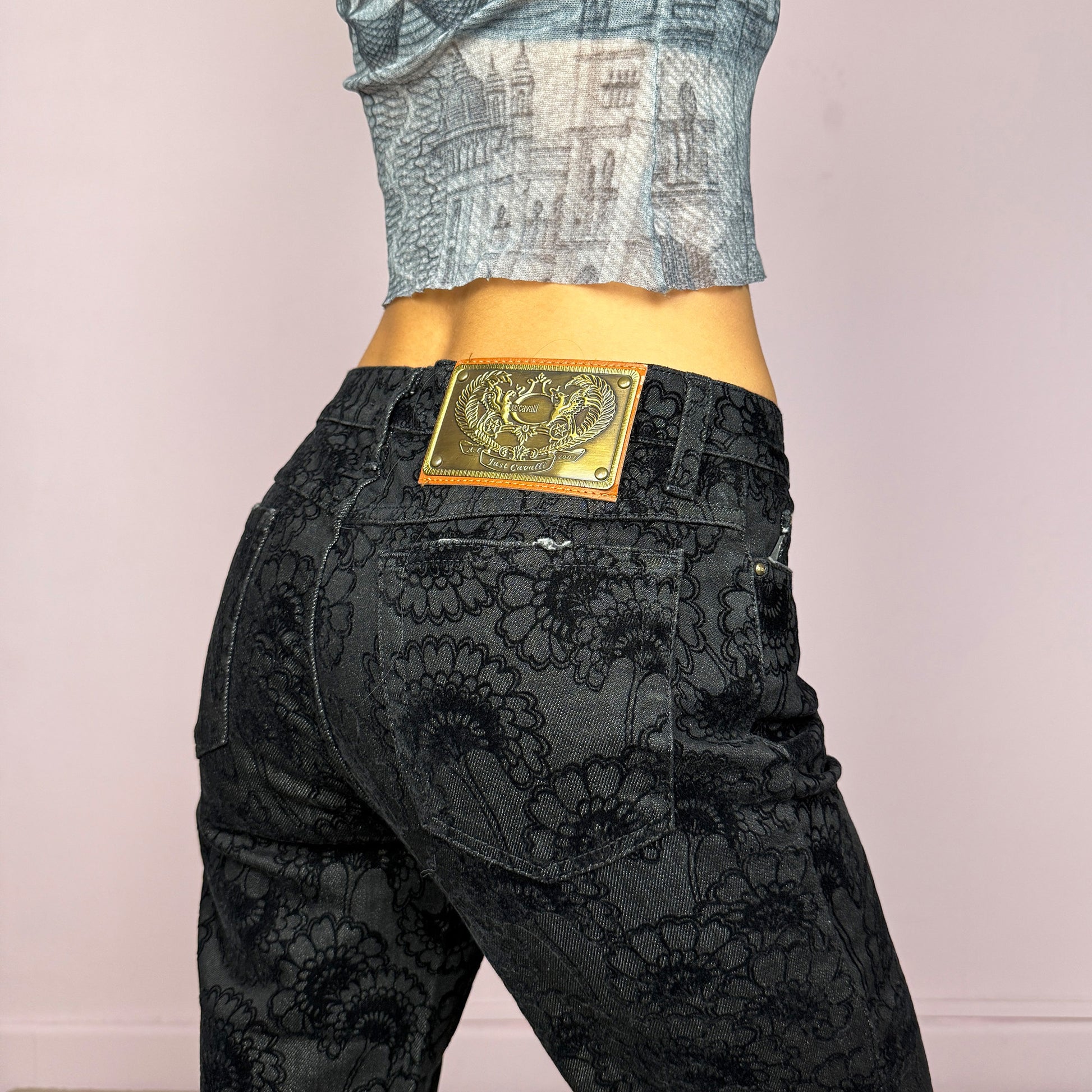 These vintage 2000s black straight jeans by Italian designer Roberto Cavalli for Just Cavalli feature a mid-rise fit and sleek straight-leg cut. Crafted from a cotton–polyester blend with slight stretch, they’re adorned with a devoré-style velvet floral pattern, blending elegance with Y2K edge. Detailed with front and back pockets, gold logo plaque, and zipper closure—ideal for night outings or elevated casual wear. Excellent vintage condition.